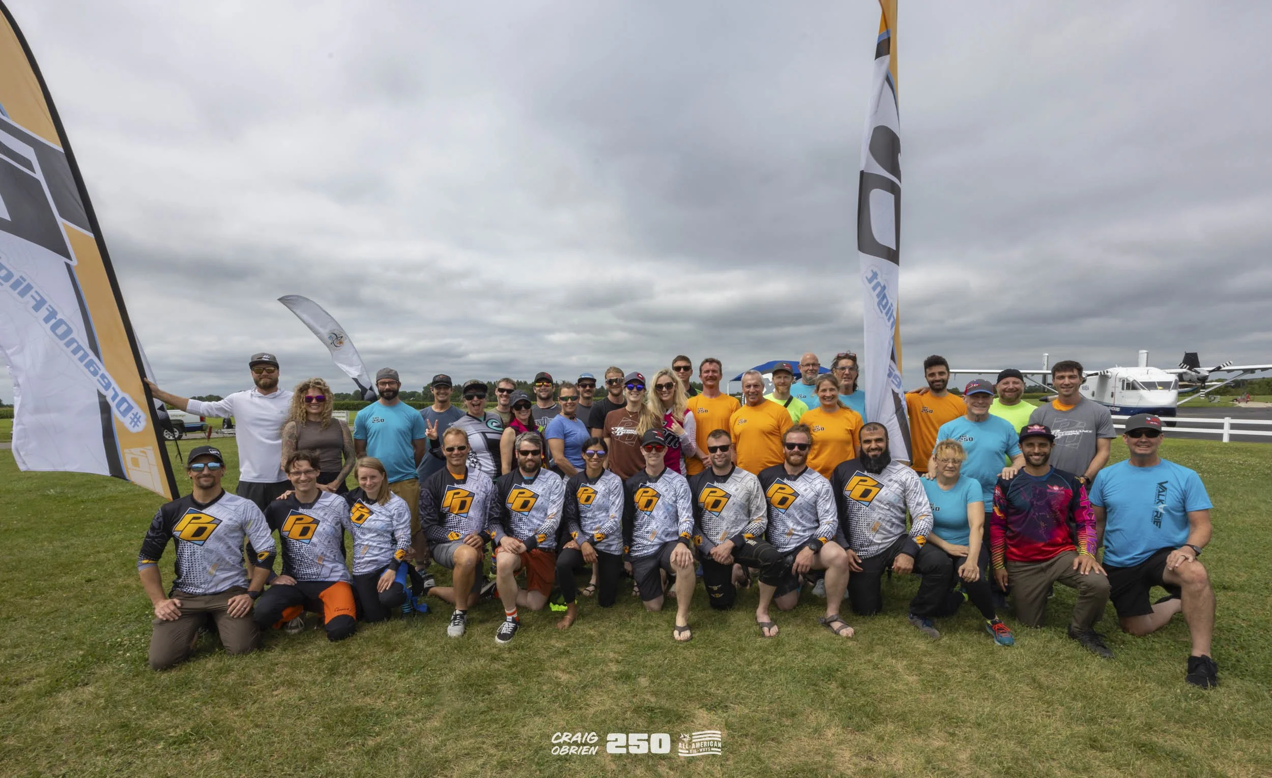 A large group of people posing outdoors on grass with a cloudy sky overhead, some wearing matching shirts and others in casual clothing. There are flags and aircrafts in the background, indicating an event or gathering related to aviation.