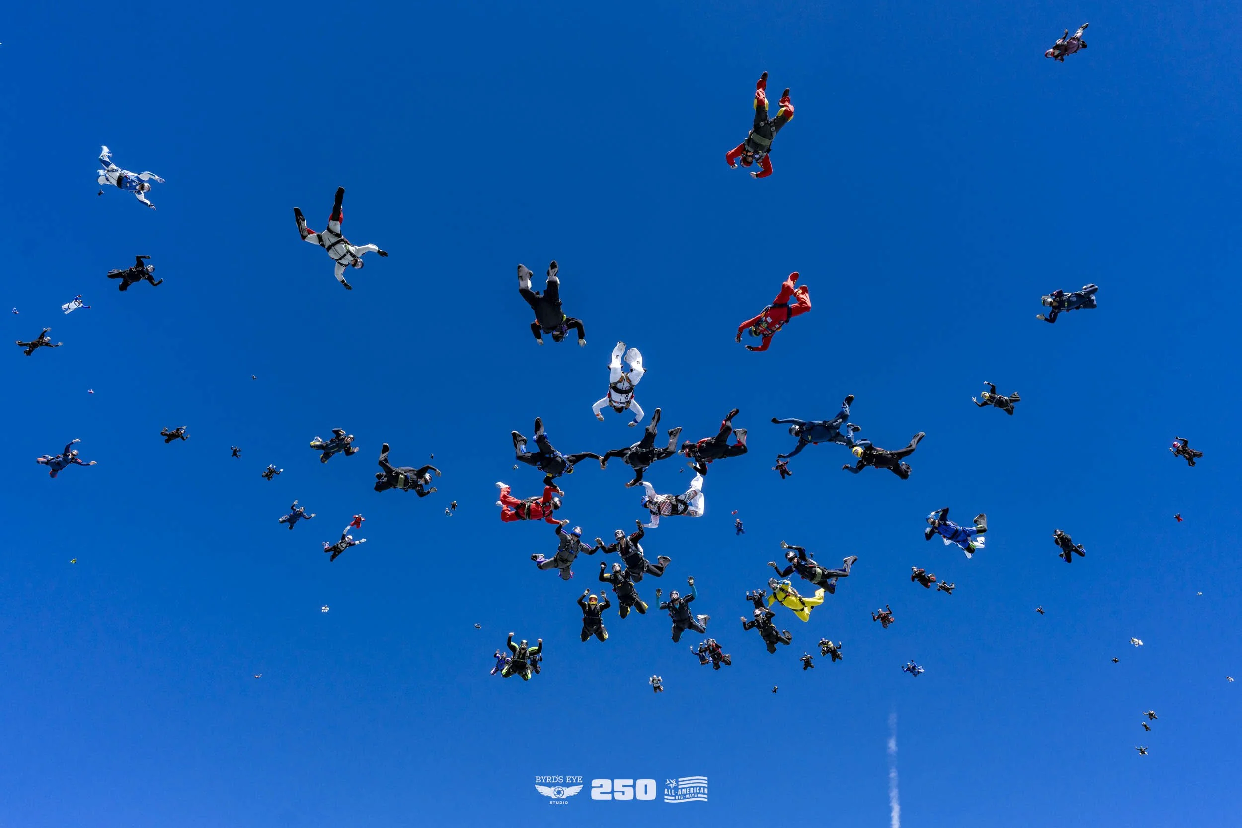A large group of skydivers wearing colorful jumpsuits and helmets, free-falling against a clear blue sky during a skydiving event.