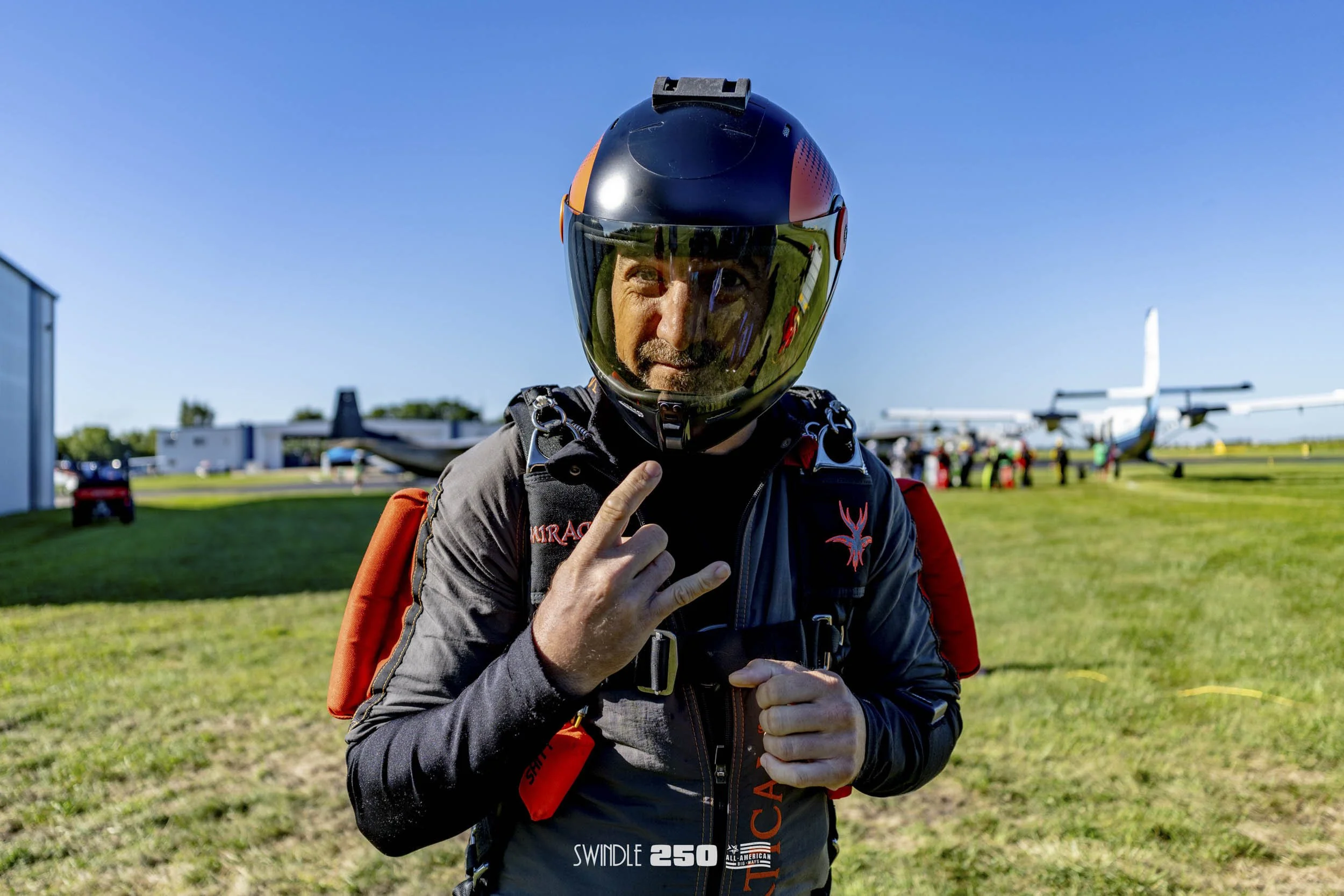 A skydiver wearing a black helmet, gray jumpsuit with red accents, and an orange backpack, making a hand sign, standing on a grassy airfield with airplanes and other skydivers in the background on a clear day.