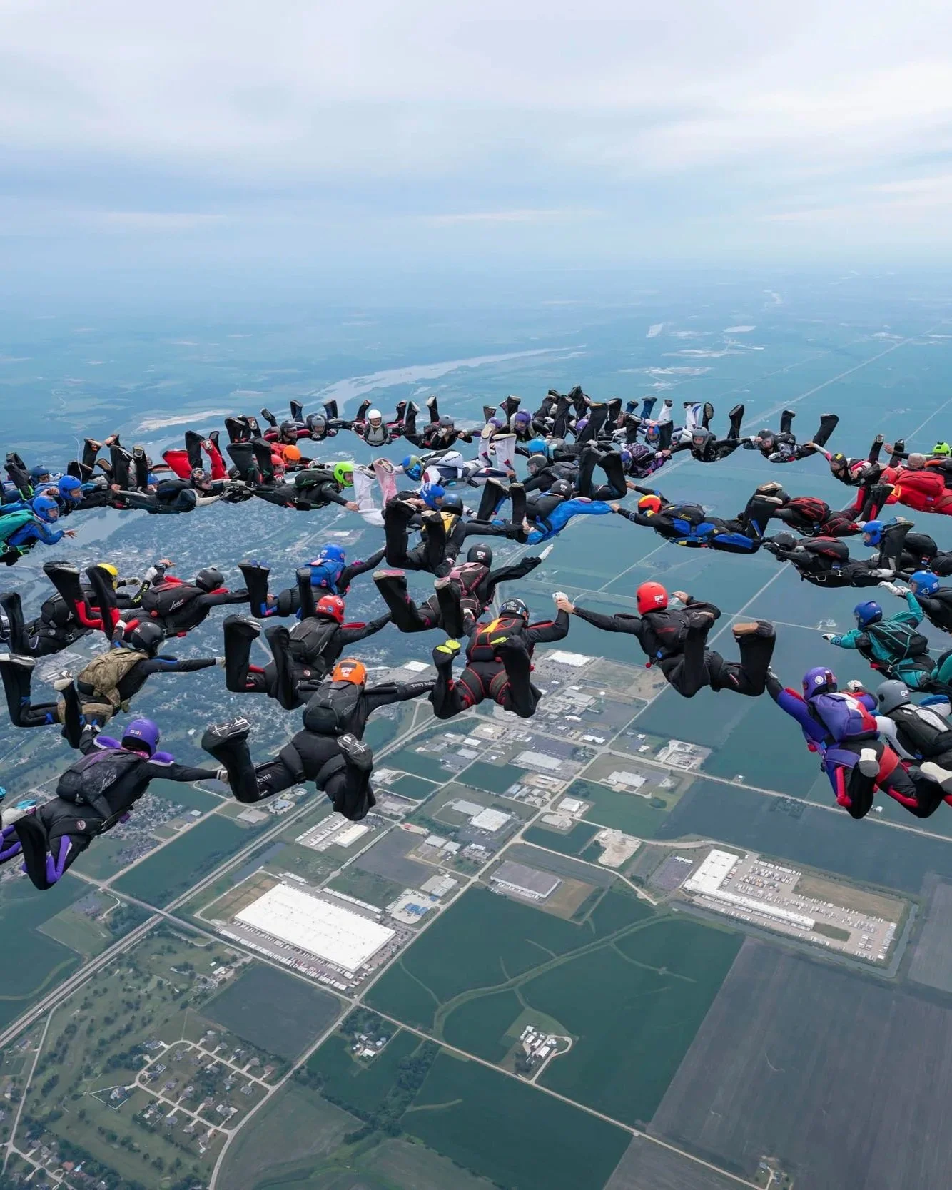 A large group of skydivers in mid-air forming a human chain above a landscape with fields and buildings.