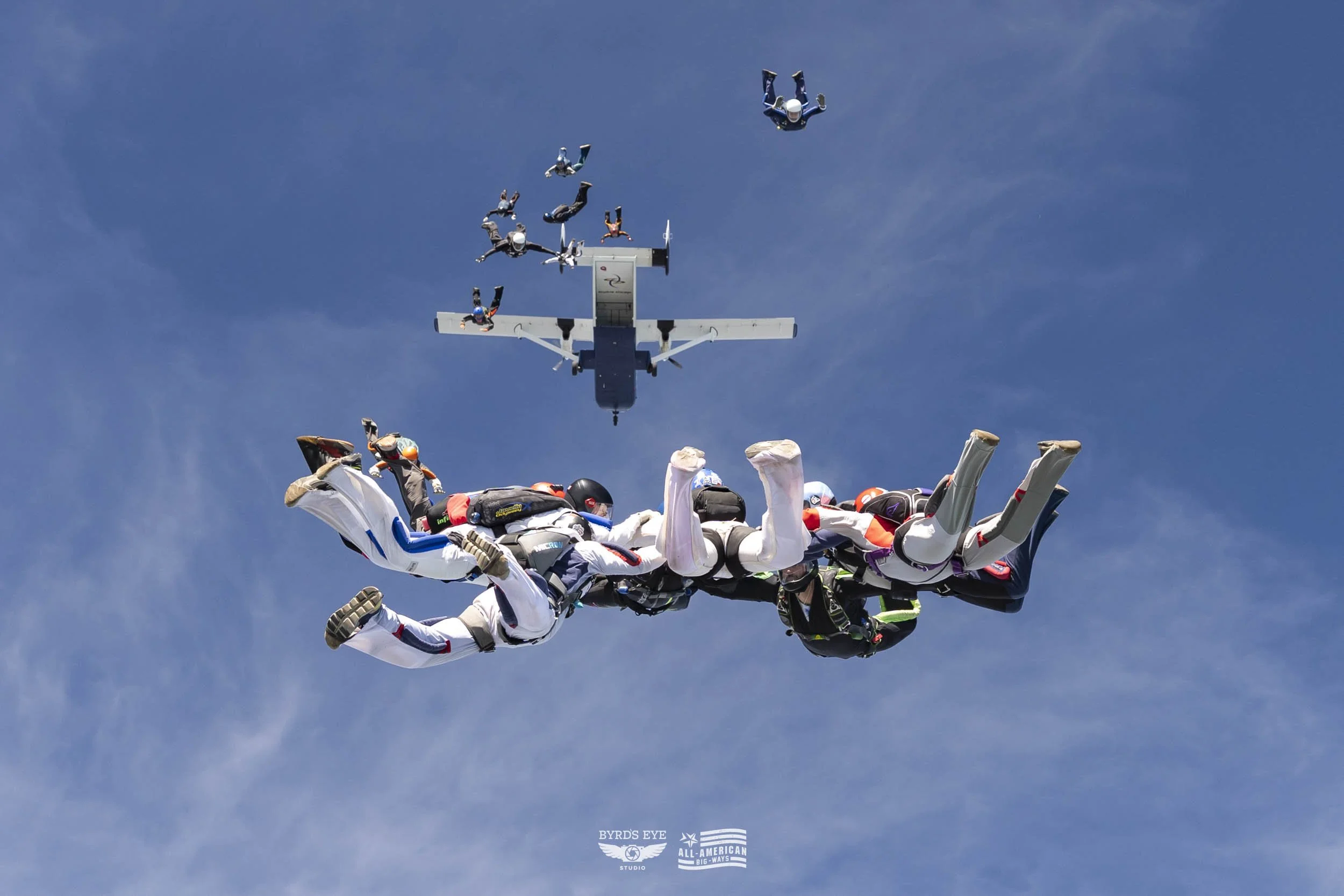 Six skydivers in free fall with an airplane in the background against a clear blue sky