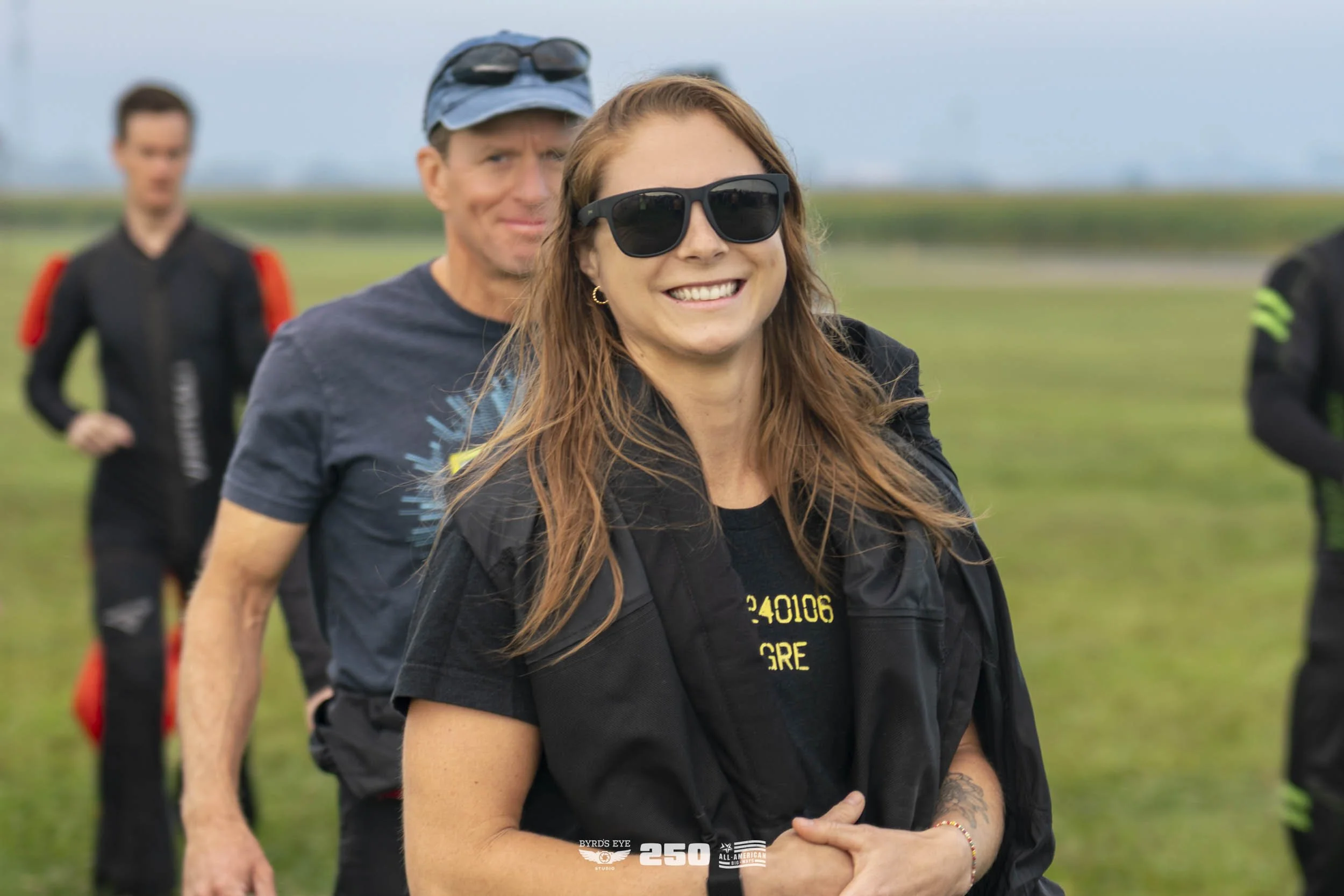 A smiling woman with long red hair wearing sunglasses, a black shirt, and a black jacket, standing outdoors on a grassy field with a group of people in the background.