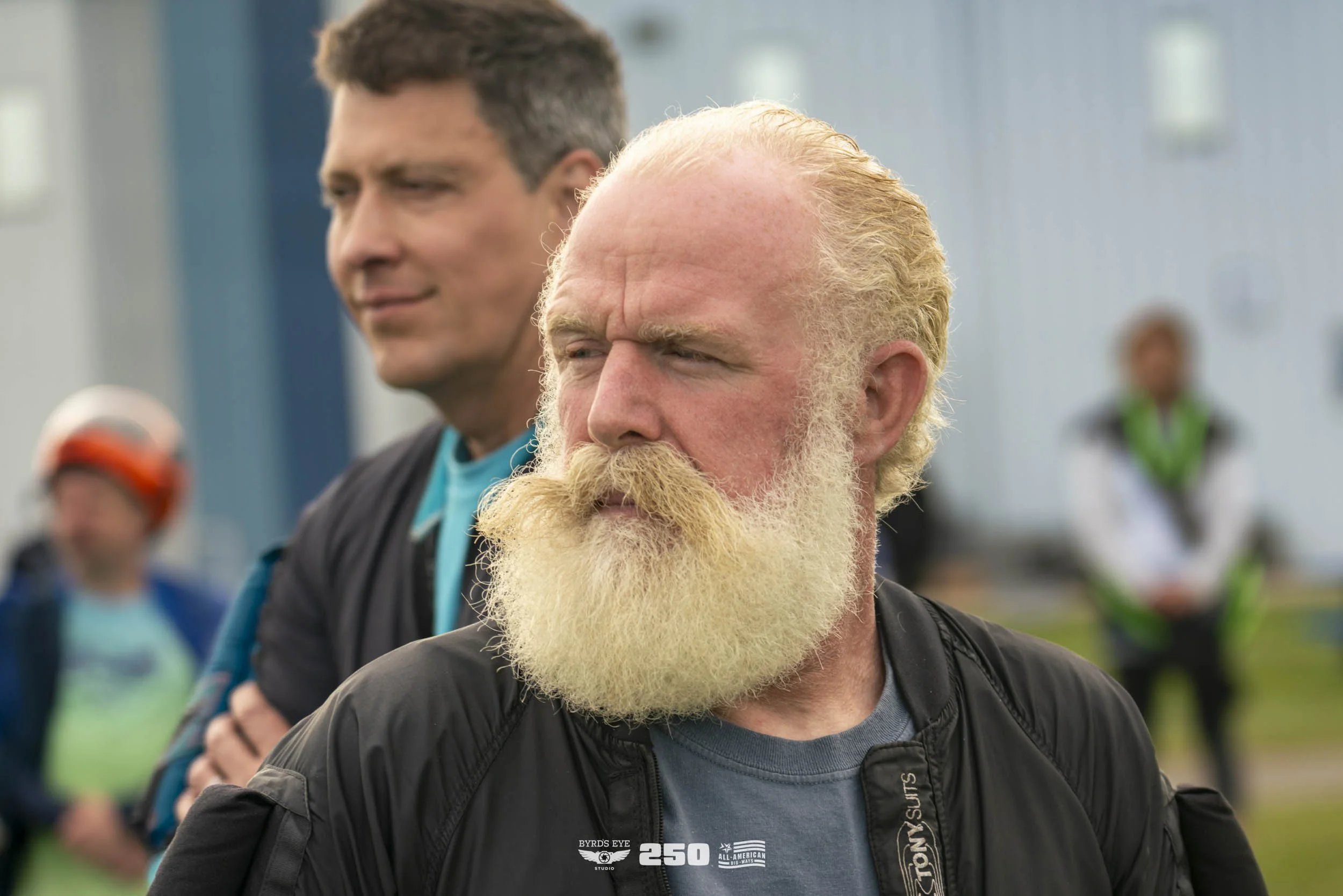A group of middle-aged men standing outdoors, with one man with a white beard and bald head in the foreground, looking serious. A man with short dark hair is slightly behind him, smirking. Other people are visible in the background, some wearing helm
