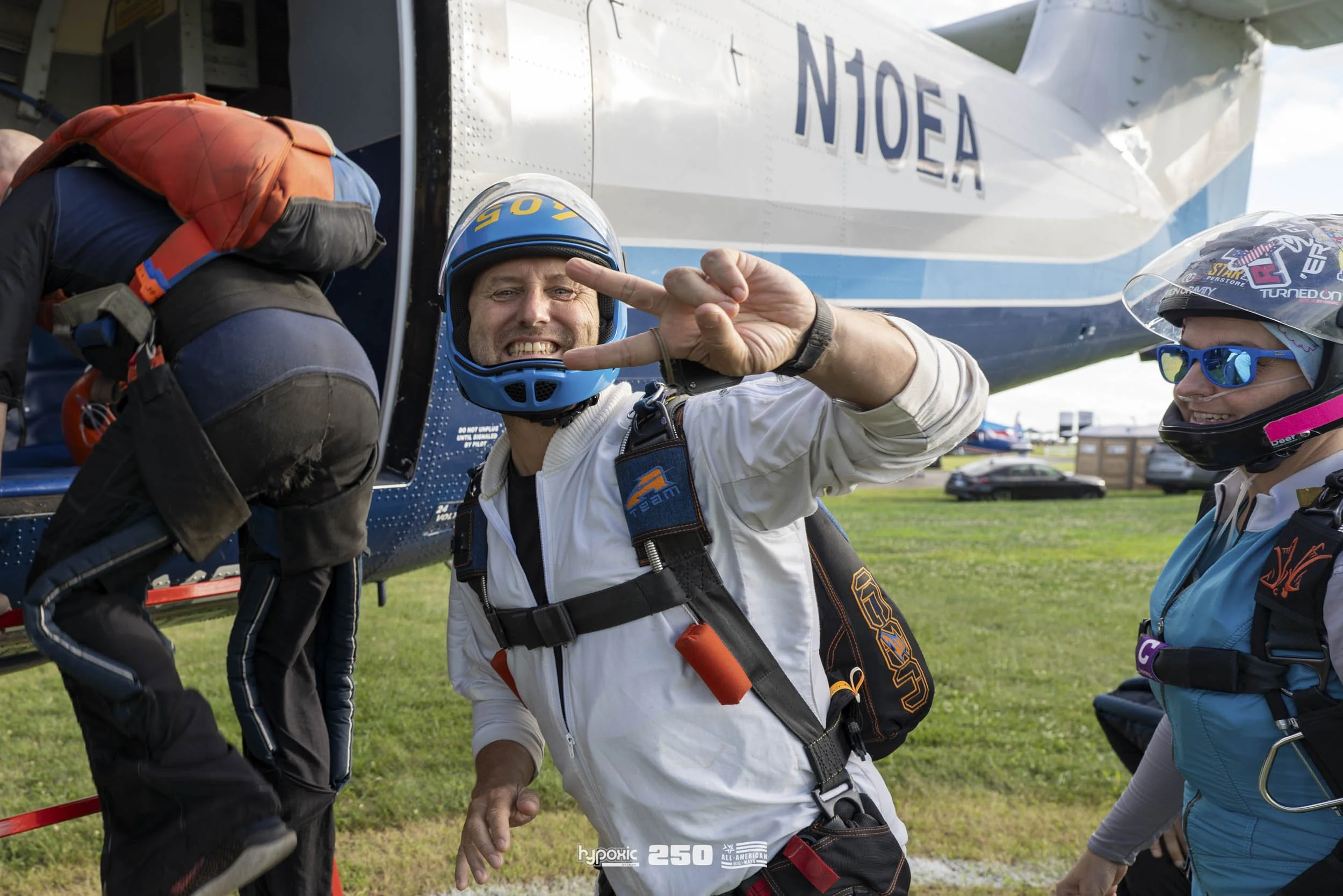 A man in a white jumpsuit and a blue helmet smiling and flashing a peace sign, standing next to an airplane with the tail number N10EA, while two other skydivers in jumpsuits and helmets are around him, preparing for a jump.