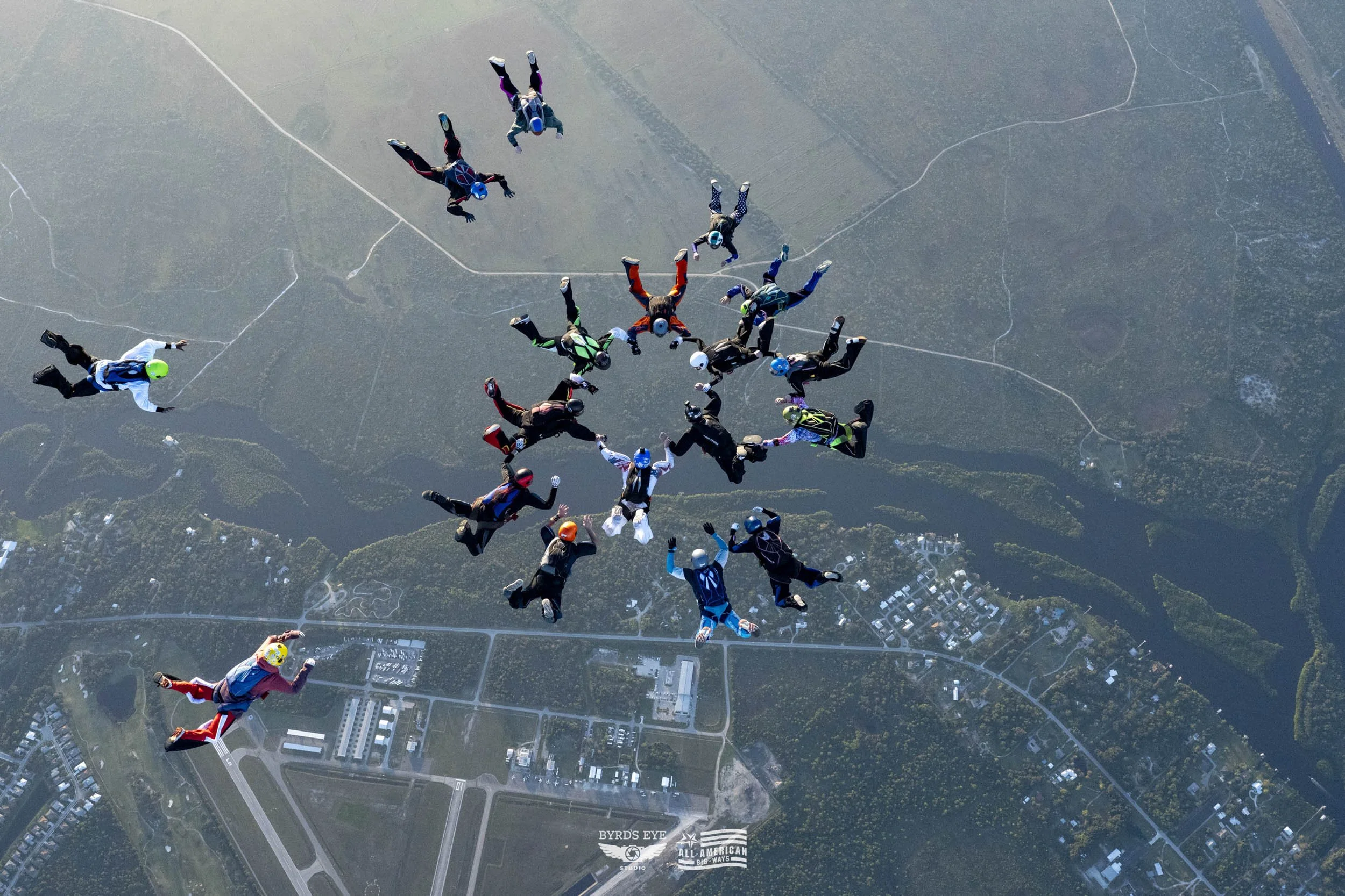 Group of skydivers in free fall above a landscape with roads, buildings, and trees.