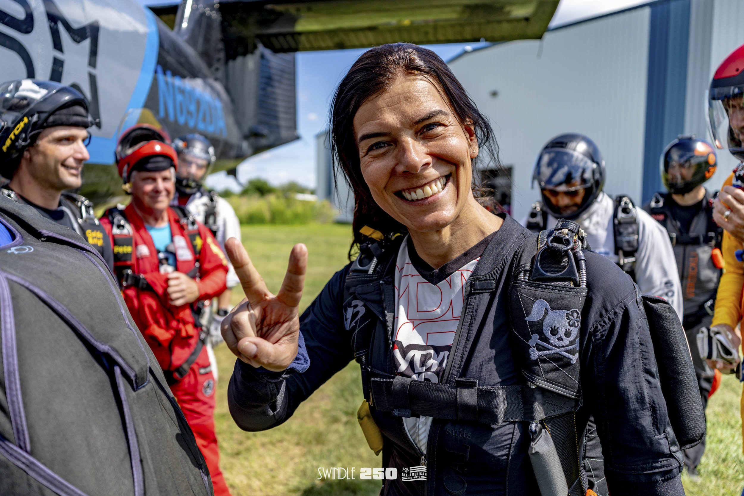 Woman smiling and making a peace sign, standing with a group of skydivers in jumpsuits and helmets, near a small plane outdoors.