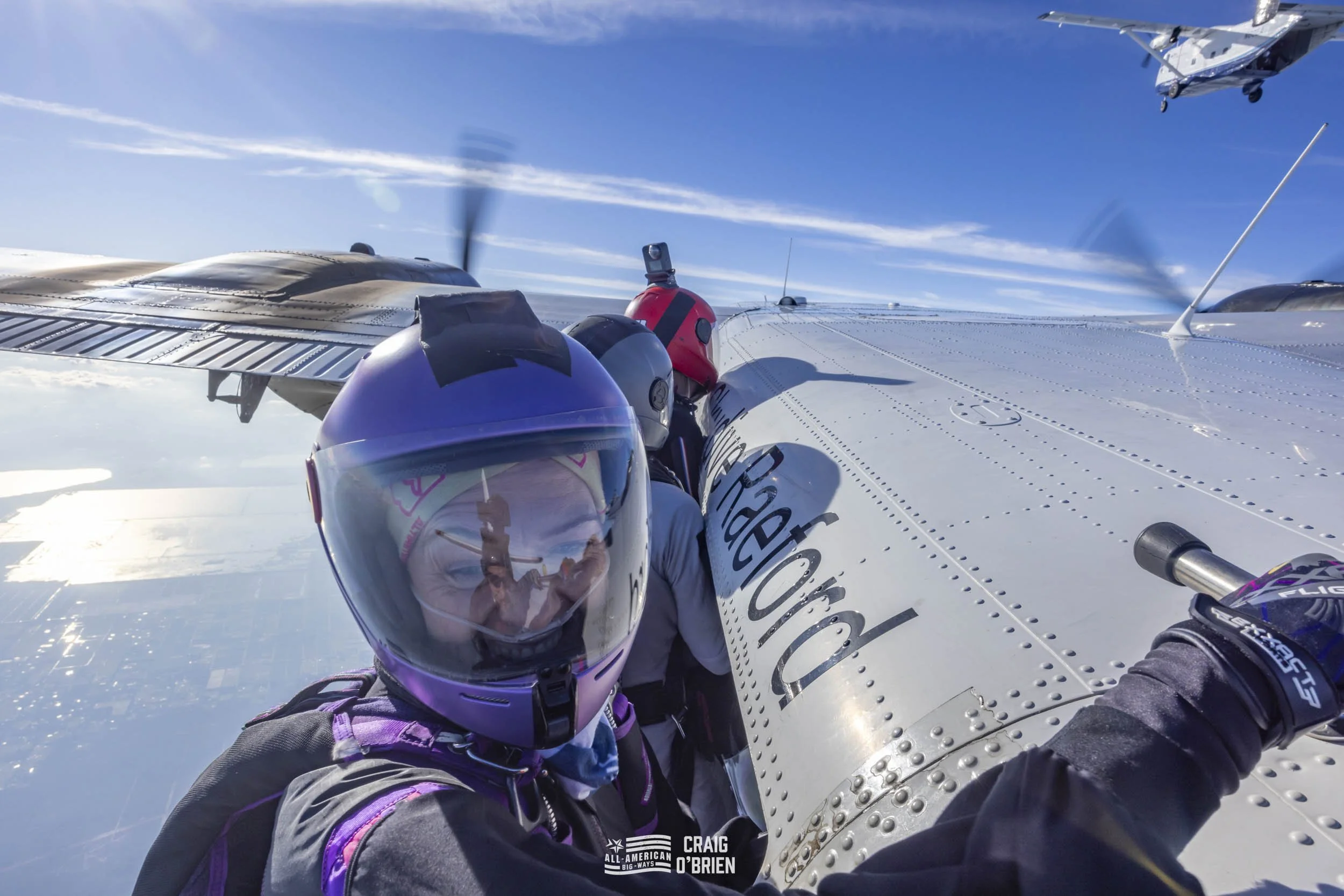 Upside-down view of a person with a purple helmet and reflective visor taking a selfie with a silver aircraft and fellow crew members, part of Team United, during an air race at high altitude under a clear blue sky.