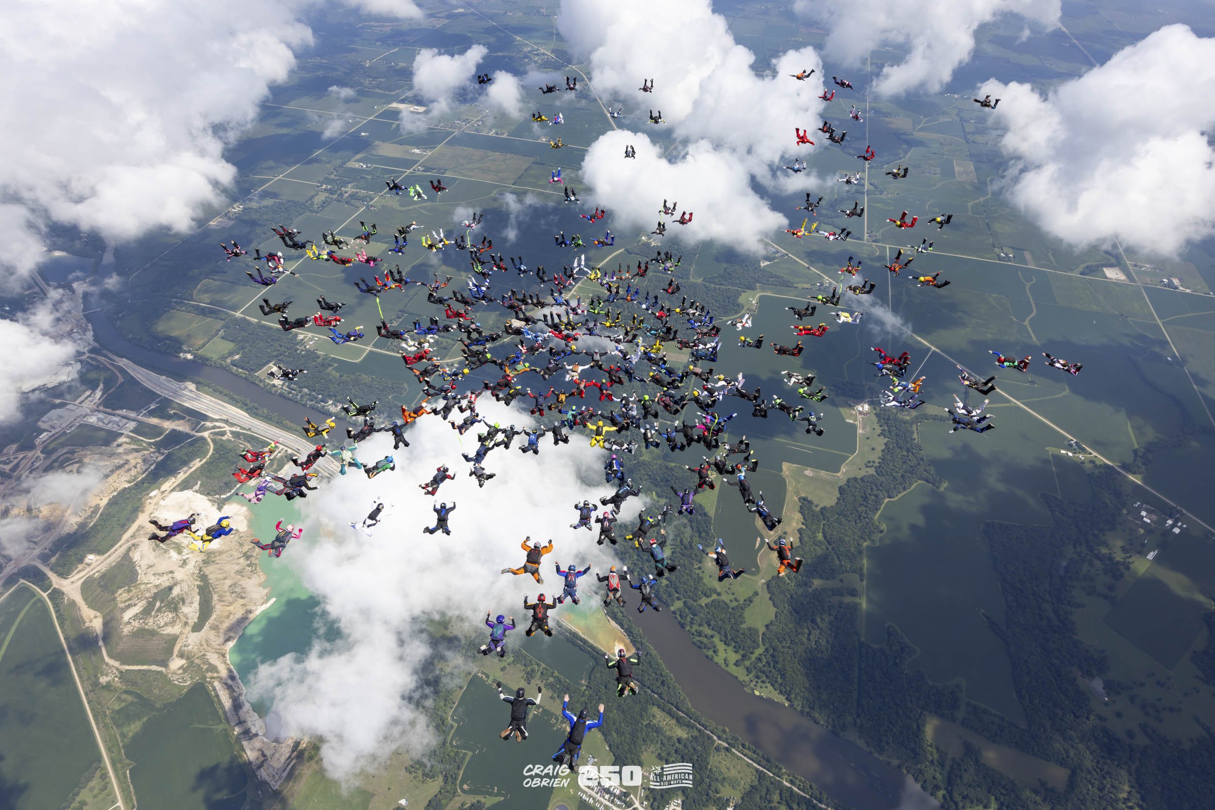 A large group of skydivers in colorful jumpsuits free-falling in formation above a landscape with water, fields, and a dam, with several clouds scattered across the sky.