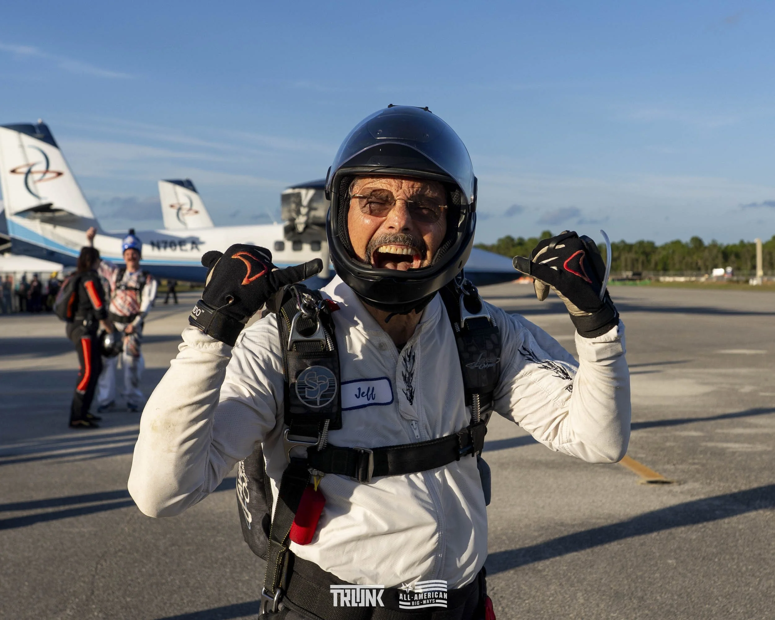A man in a pilot uniform, wearing a helmet and gloves, smiling and pointing to his head while standing on an airport tarmac with commercial airplanes in the background.