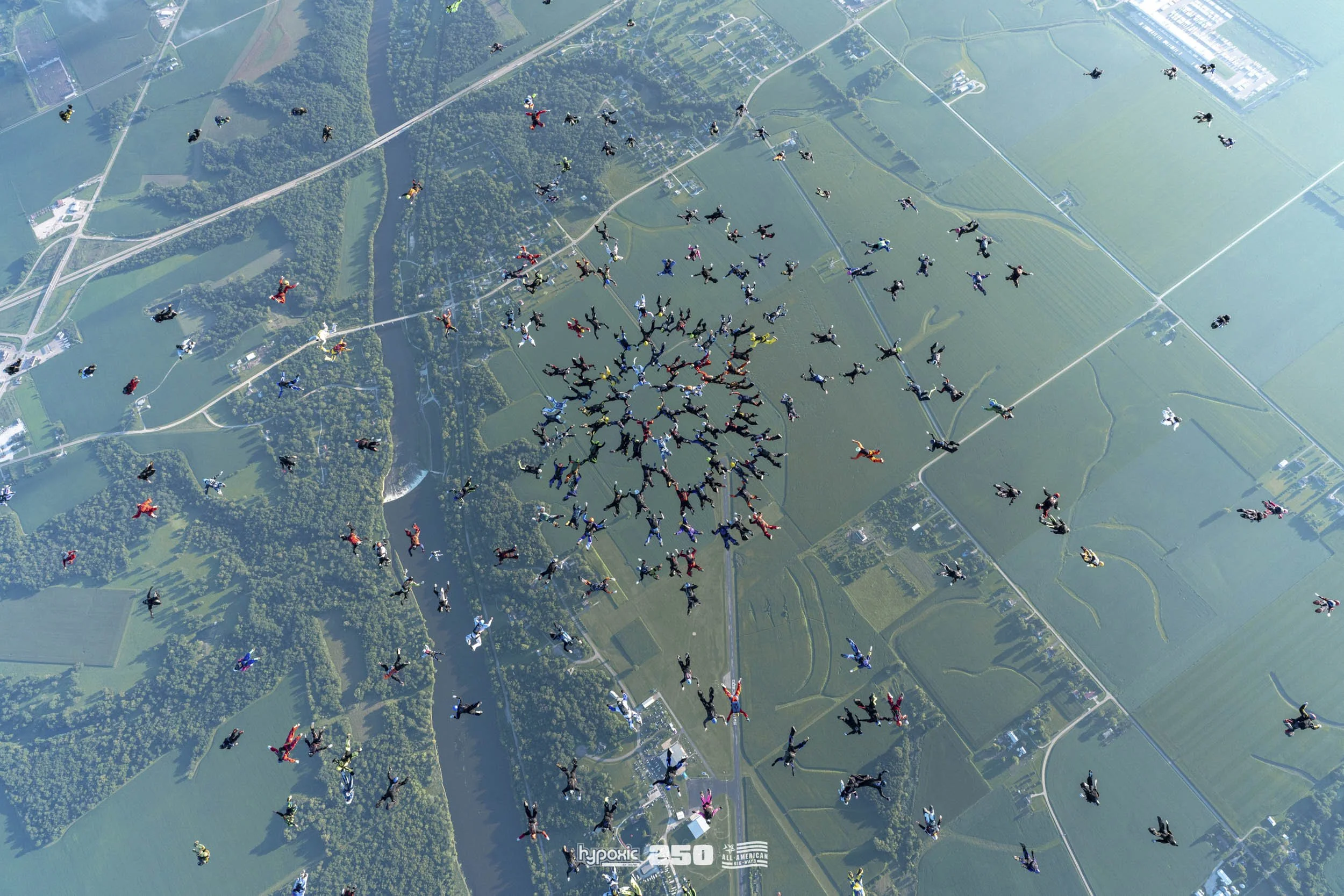 Aerial view of hundreds of skydivers in a formation above open farmland with green fields, trees, and roads.