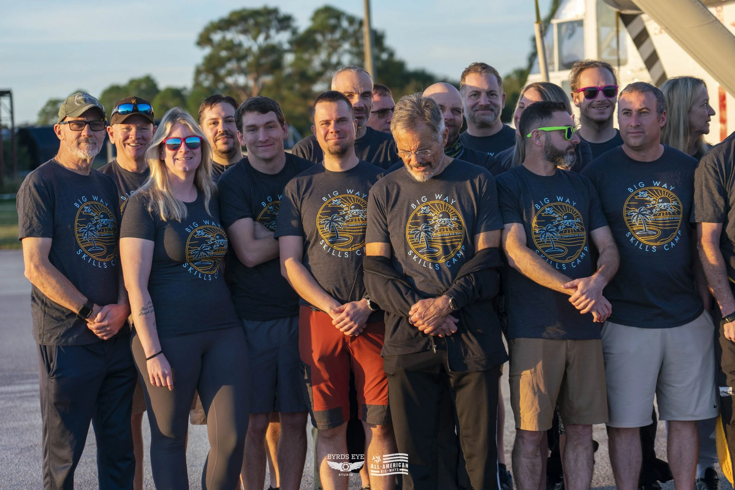 Group of people wearing matching dark blue t-shirts with a tropical sunset design, standing outdoors during sunset, some wearing sunglasses, smiling and looking towards the camera.