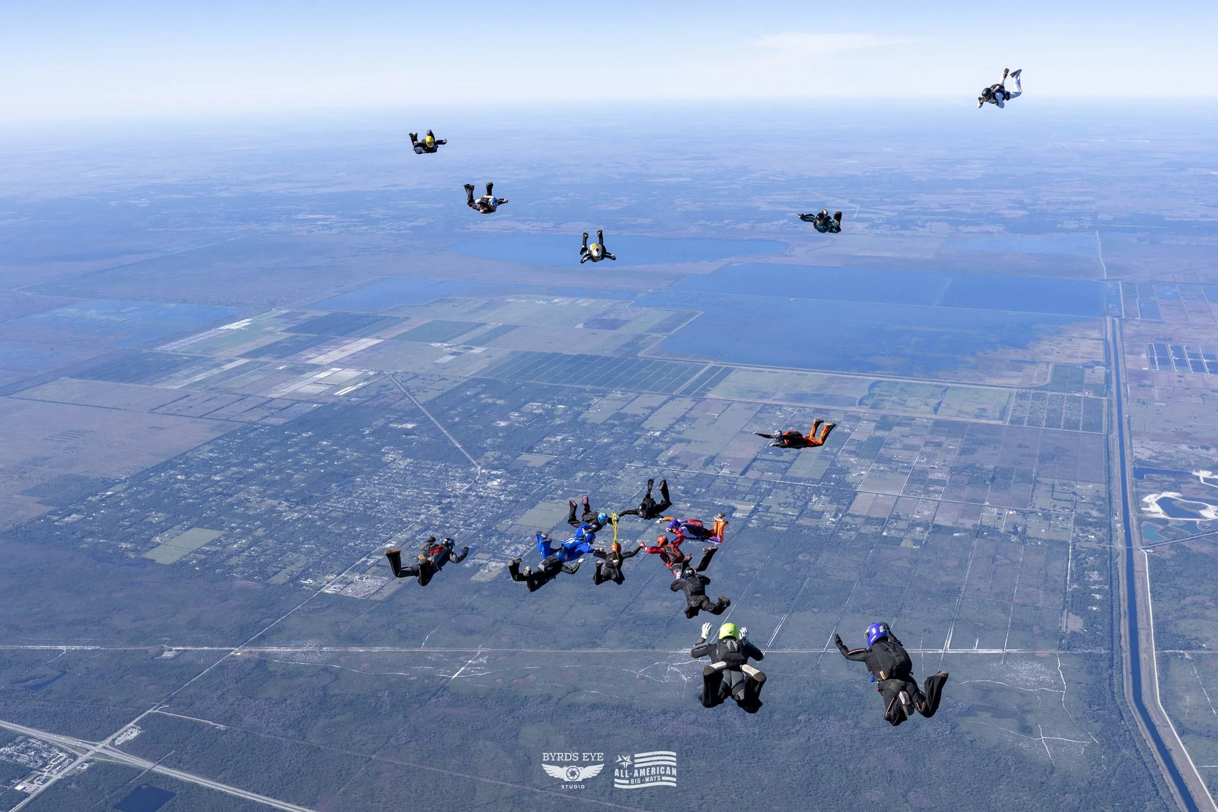 Group of skydivers free-falling over a landscape with fields and a river, seen from above.