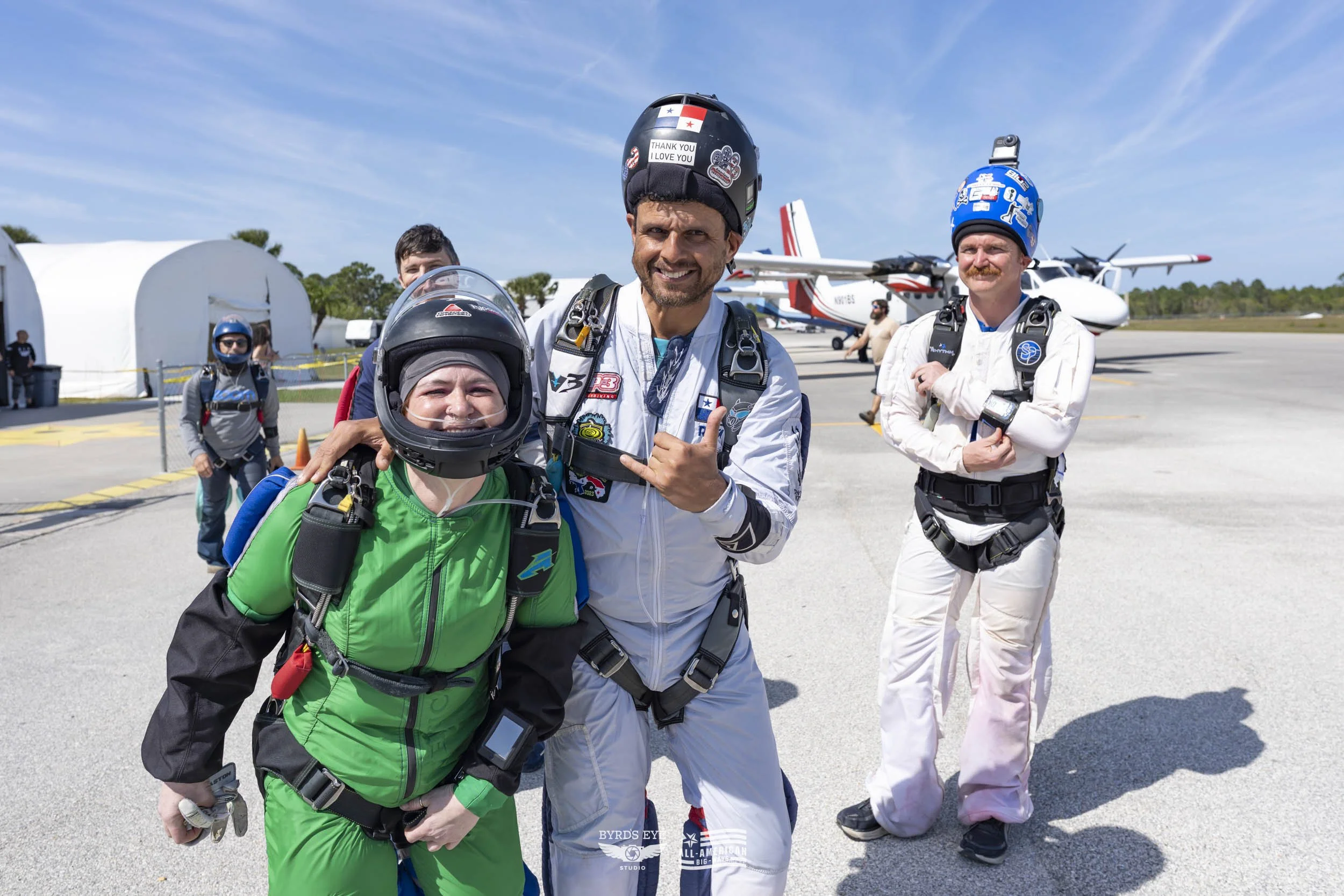 Skydivers in jumpsuits and helmets posing on an airfield with aircraft in the background, some members smiling and making hand gestures.