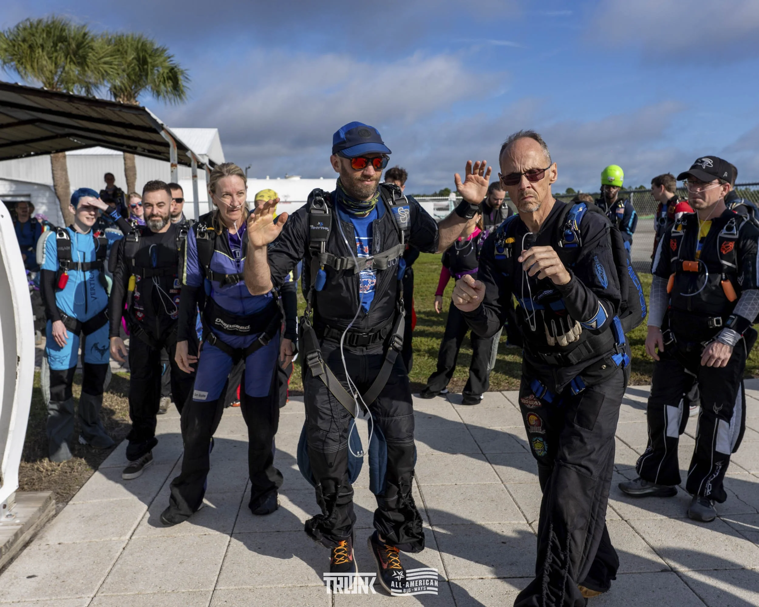 A group of skydivers dressed in jumpsuits and harnesses standing together outdoors, preparing for a skydiving jump under a partly cloudy sky.