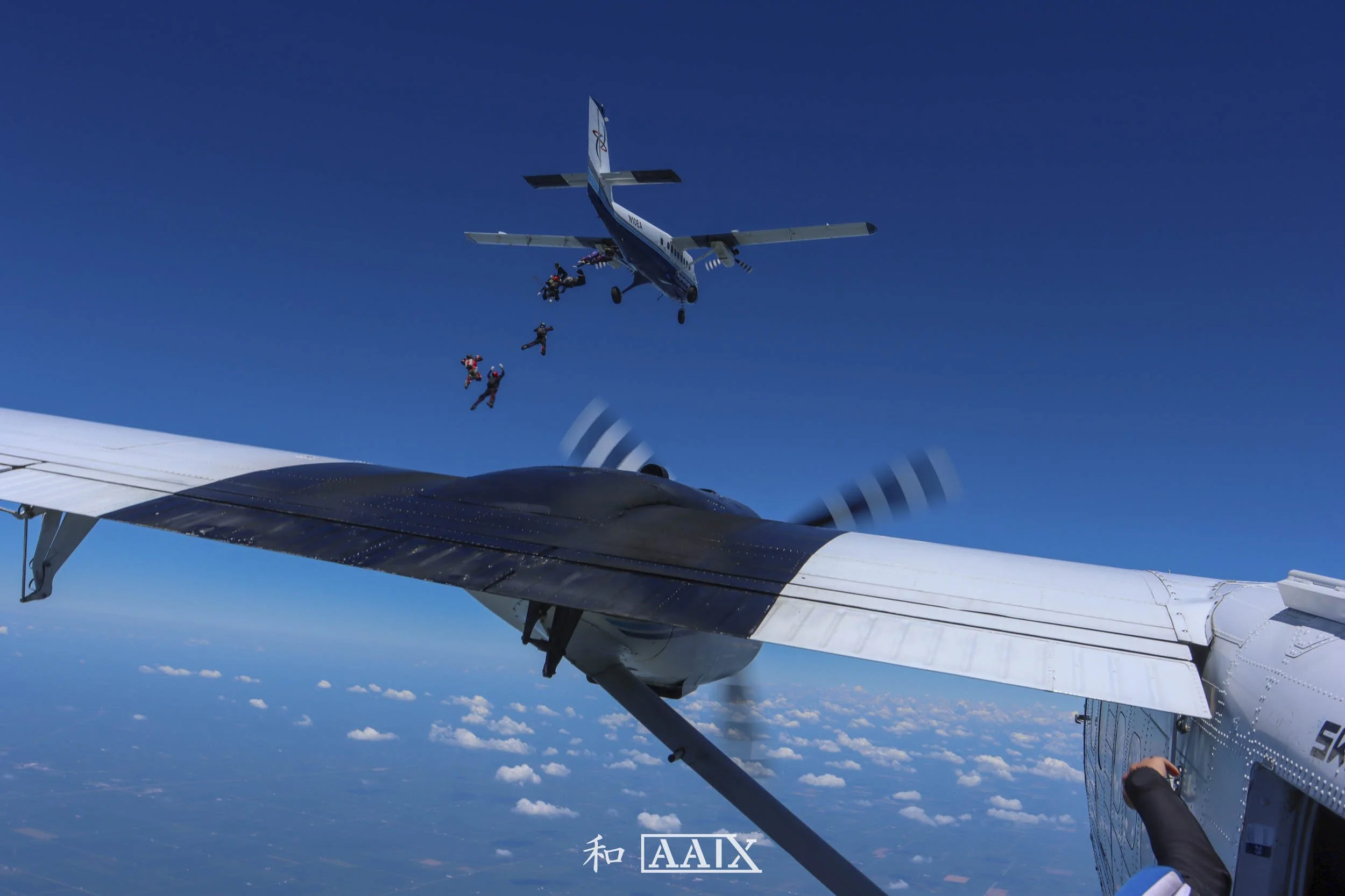 Skydivers jumping from a small aircraft in flight, with a large drone or aircraft nearby against a blue sky and scattered clouds.