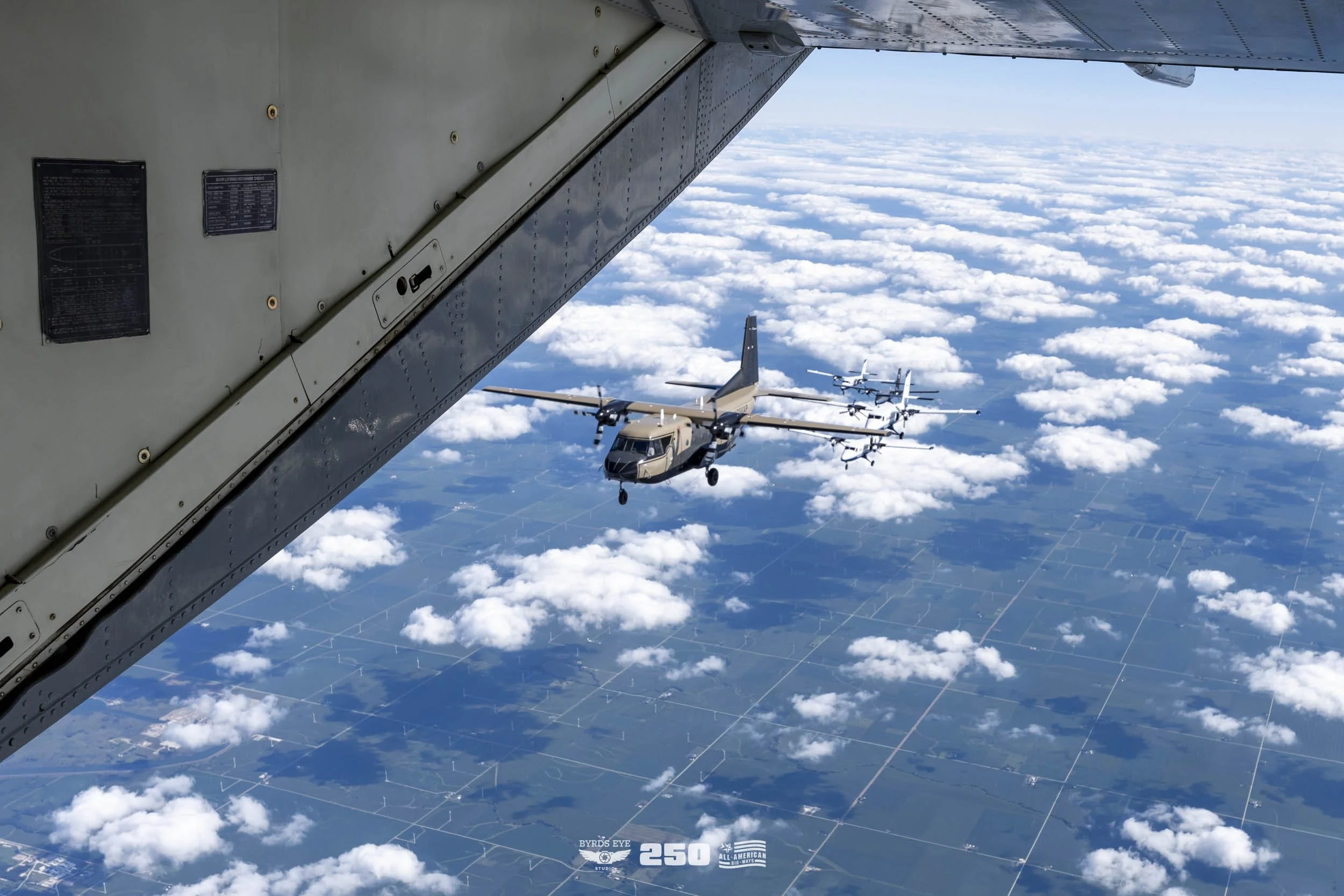 View from an aircraft window showing a military transport airplane flying above a patchwork of farmland with clouds in the sky.