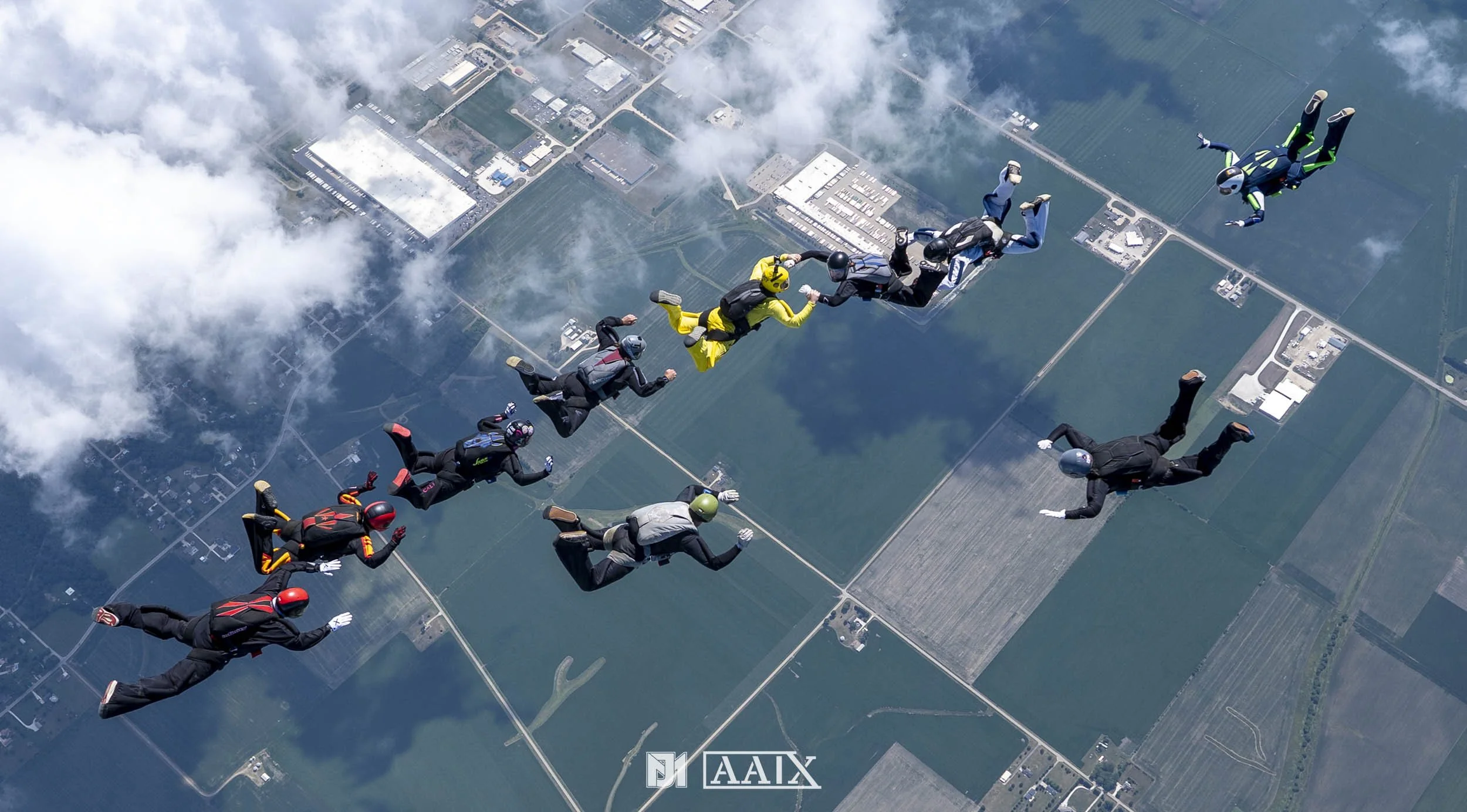 A group of skydivers in freefall formation above a landscape with fields and buildings, holding hands in a circle.