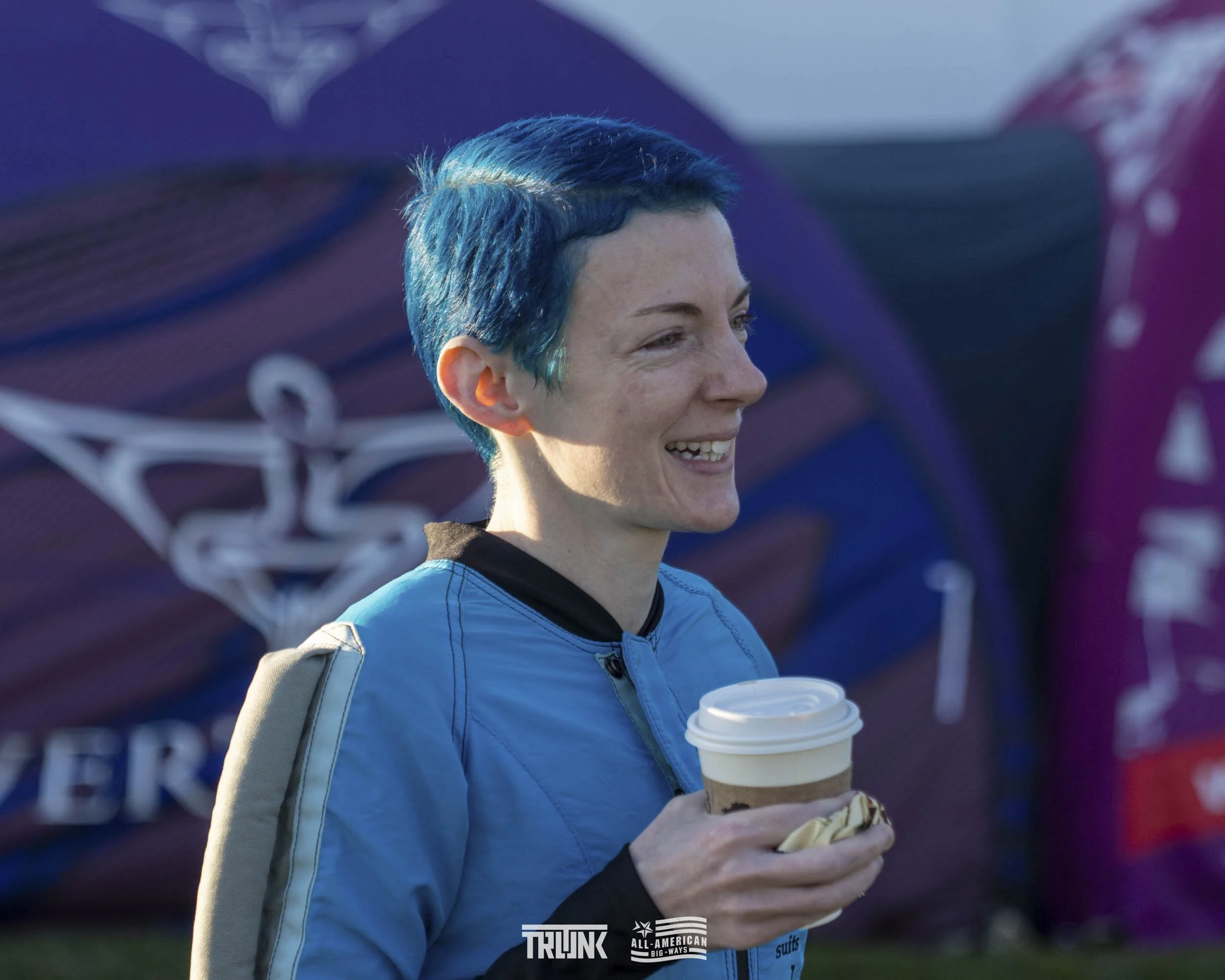 Smiling woman with short blue hair holding a coffee cup and an ice cream cone, outdoors with banners and a blue sky in the background.
