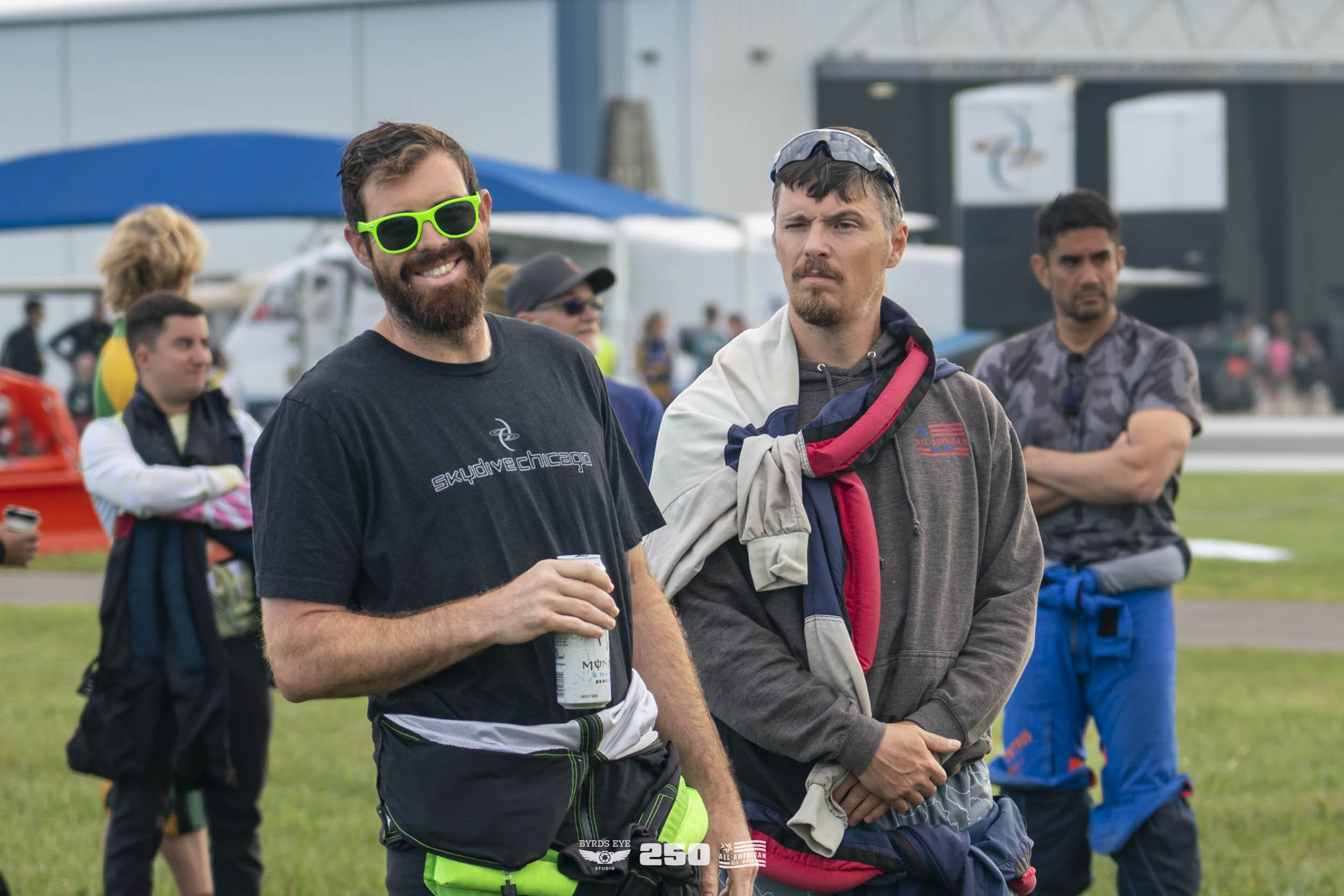 Two men standing outdoors at an event, one smiling wearing sunglasses and holding a drink, the other with a serious expression, wearing a hoodie with a jacket draped over shoulders. Several other people are in the background on a grassy area.