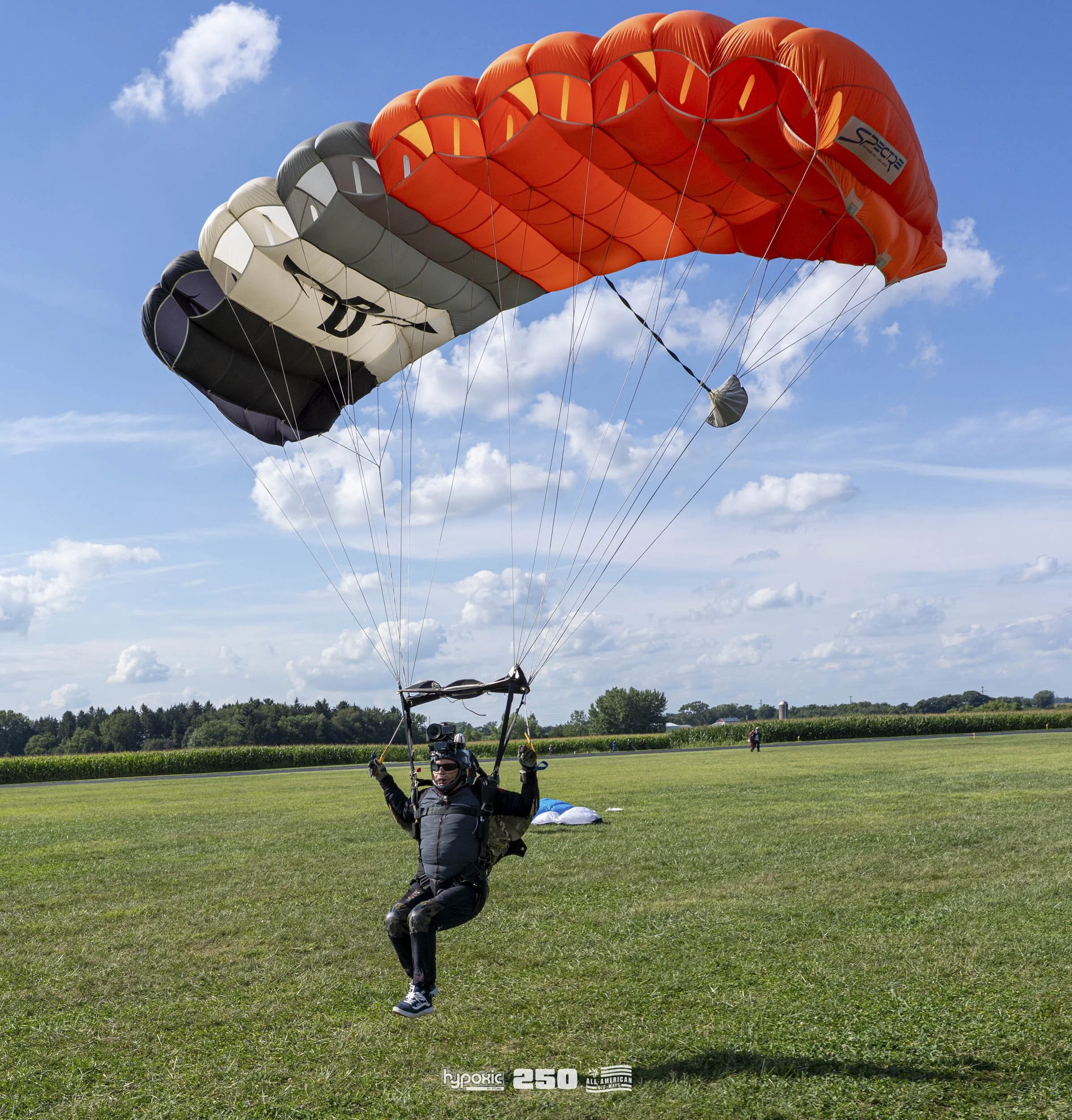 A person in a black parachuting suit is landing on a grassy field with a parachute that is red, black, gray, and white. The sky is partly cloudy with some blue visible.