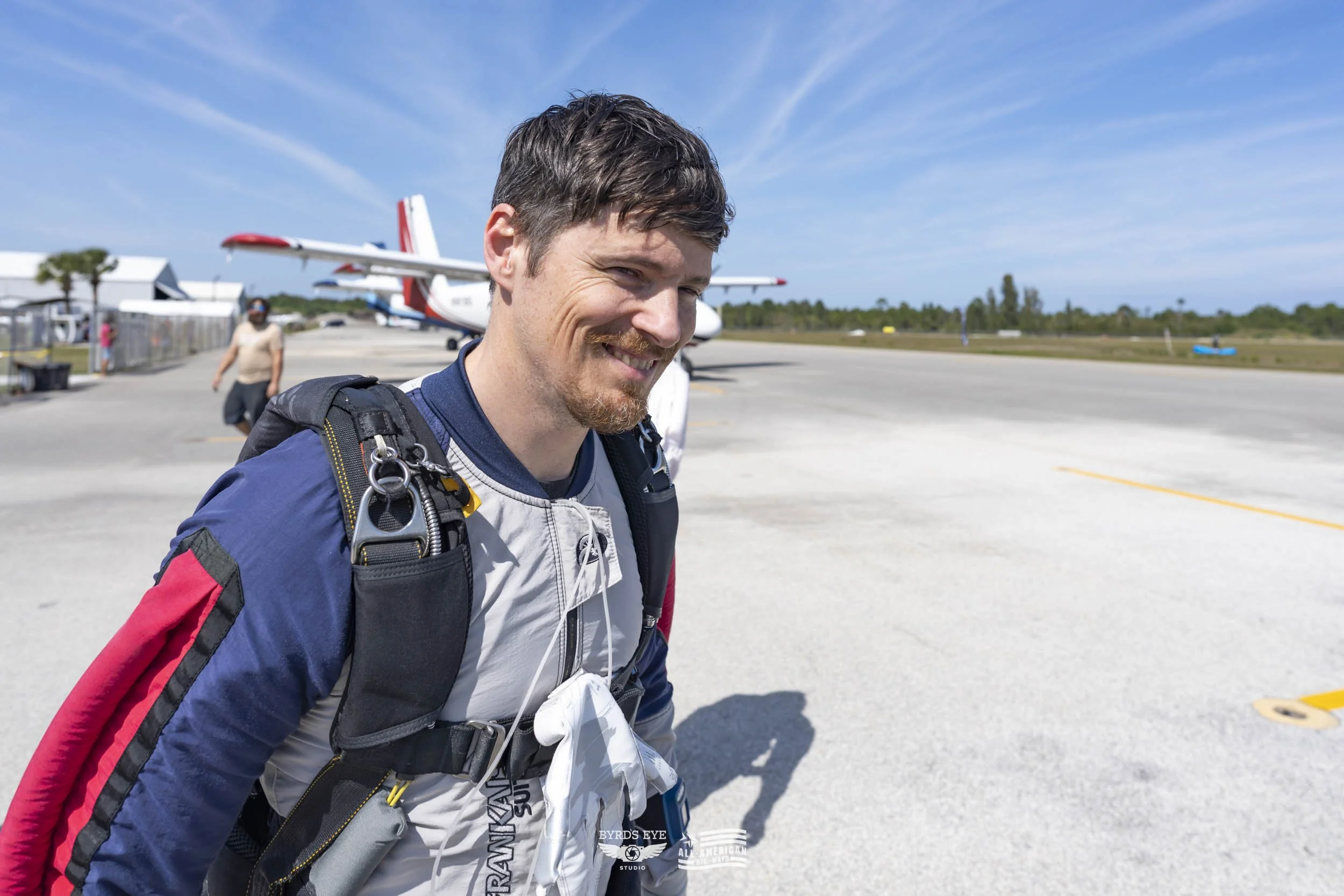 A man with a backpack smiling at an airport tarmac with planes in the background on a sunny day.