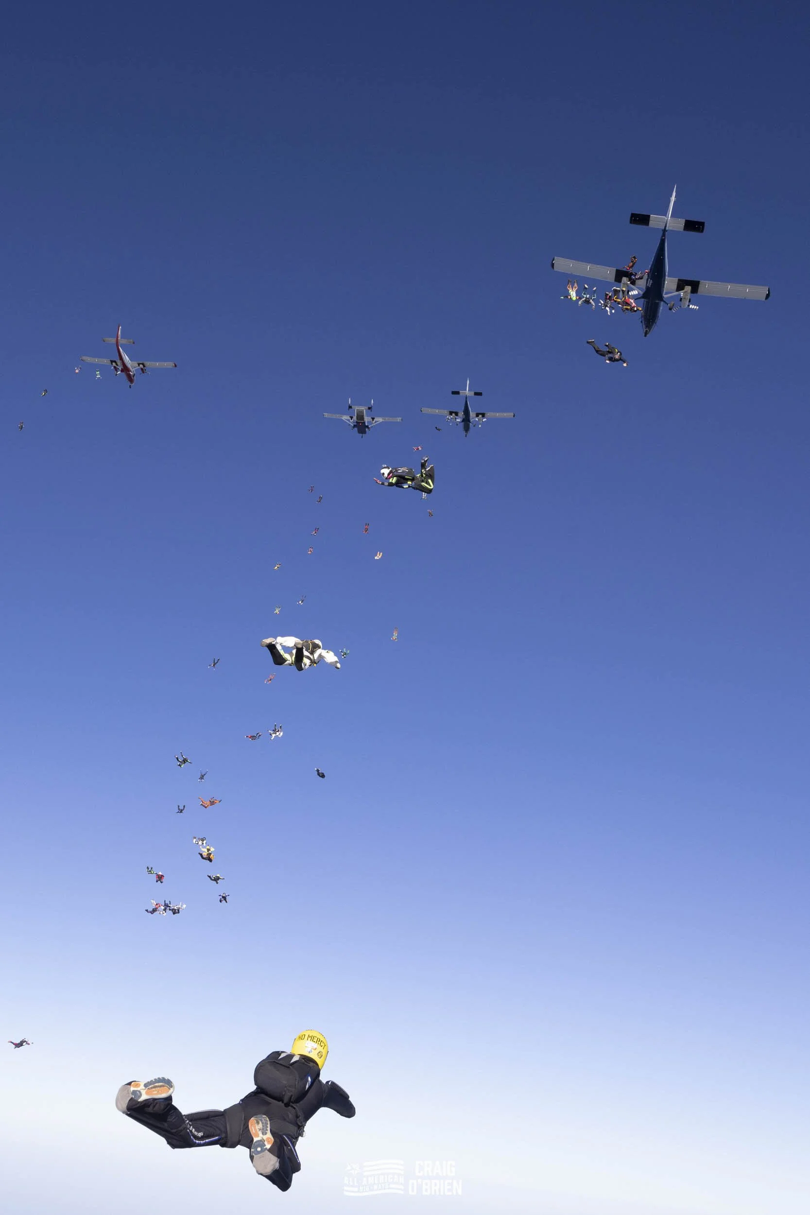 Skydivers in free fall formation with a clear blue sky background, some in tandem and others with individual gear, at a large altitude.
