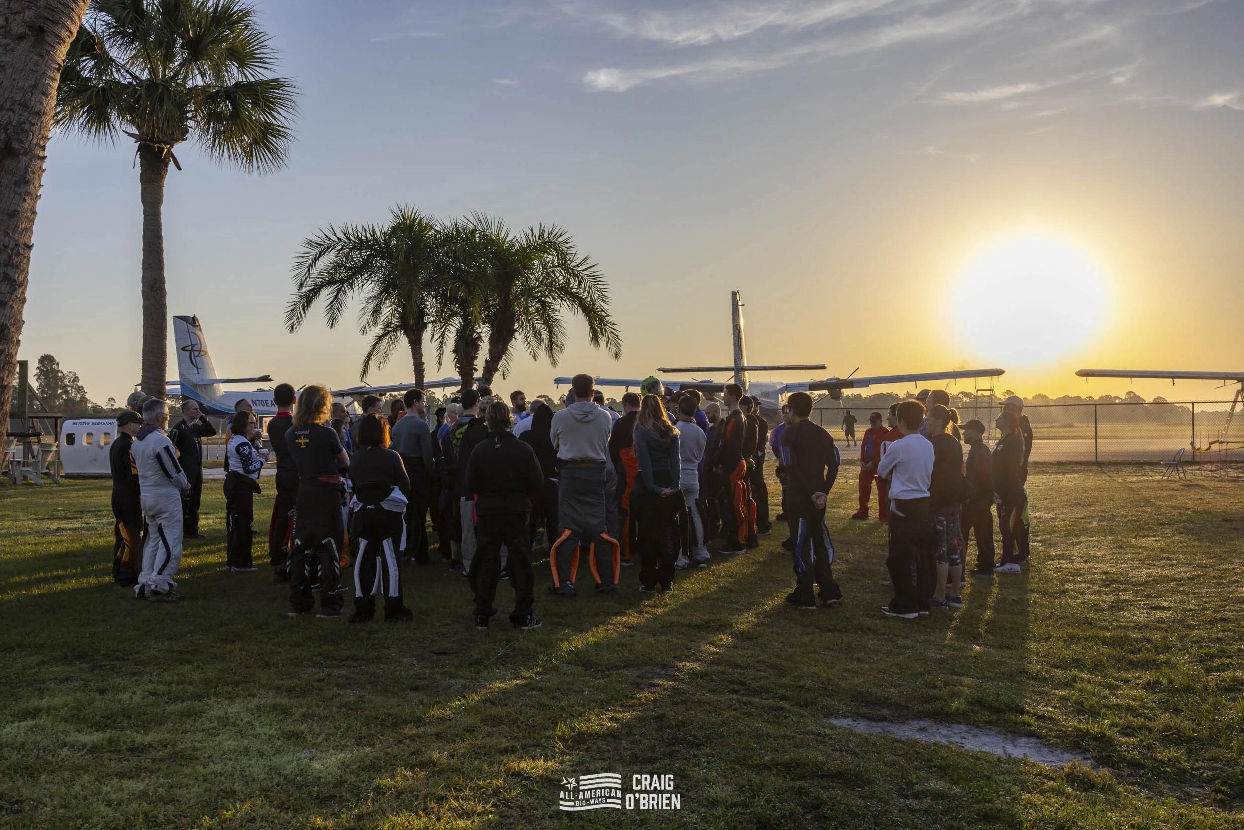 A group of skydivers gathered outdoors during sunrise near small aircraft, with palm trees and a chain-link fence in the background.