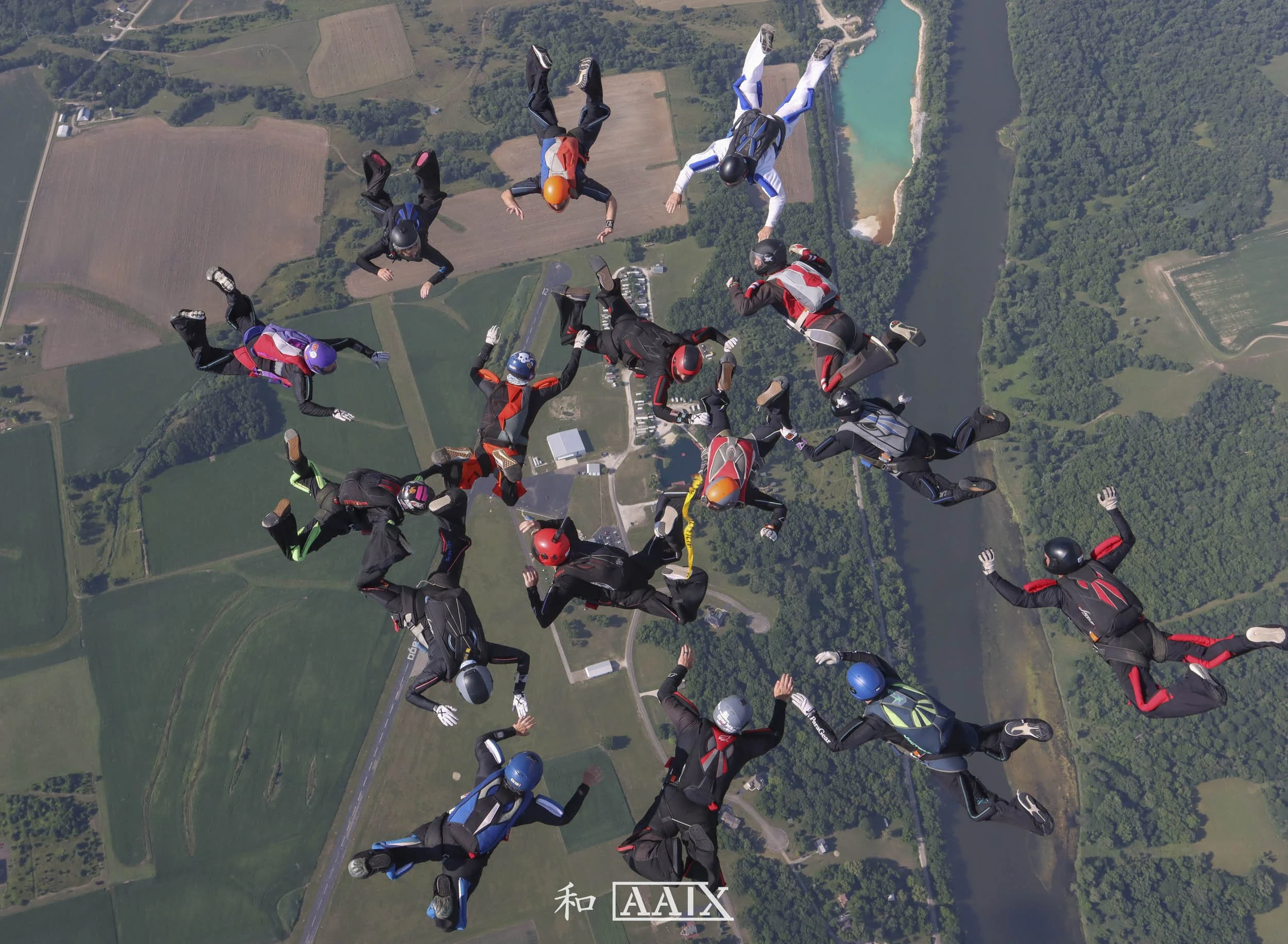 A group of skydivers in free fall over a rural landscape with fields, trees, a river, and some buildings.