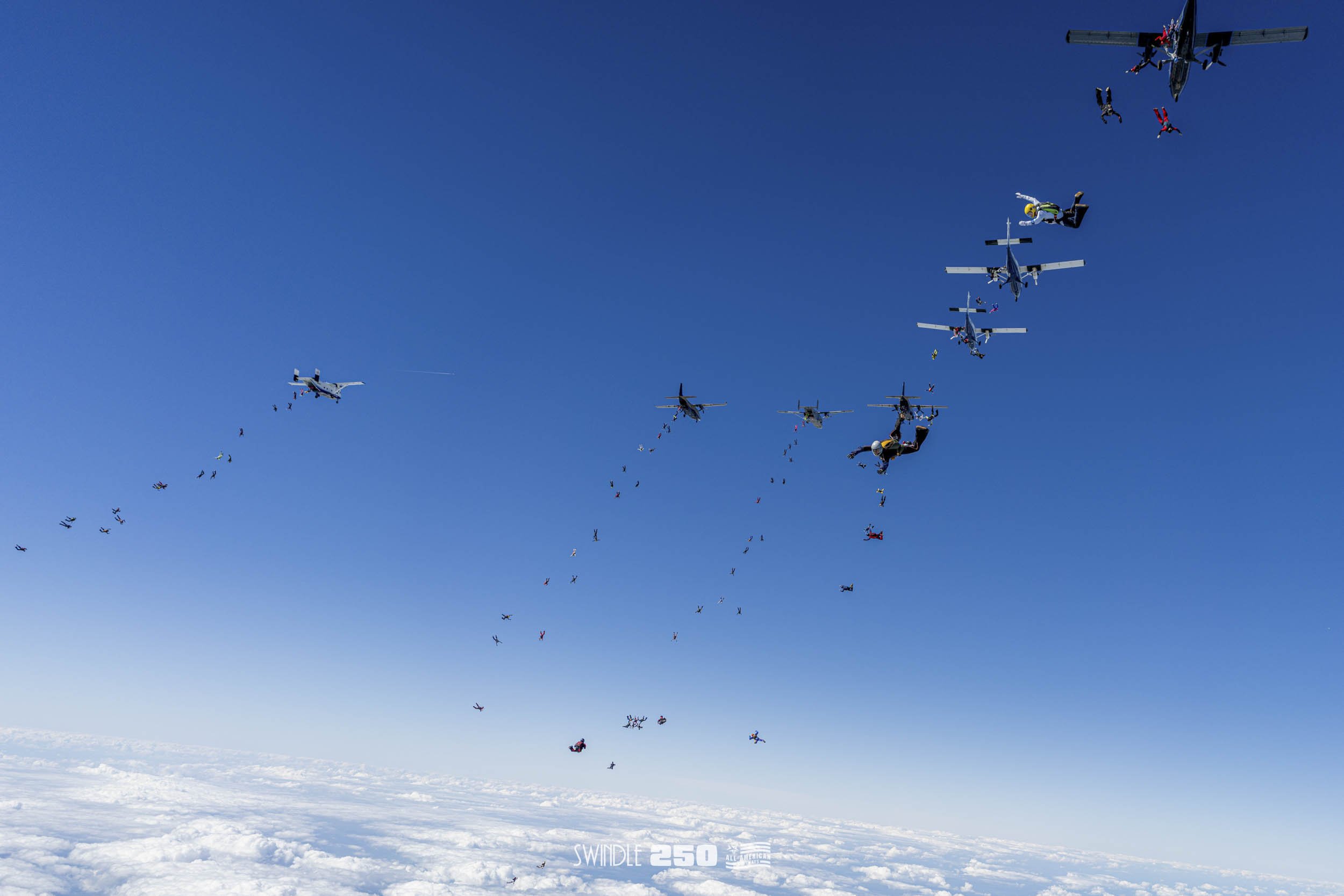 Skydivers jumping from airplanes in formation over a cloudy sky with some parachutes deployed.
