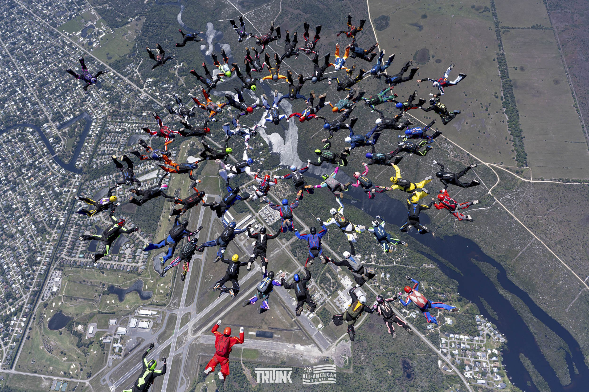 Skydivers forming the shape of a tree in freefall over a city and park area.