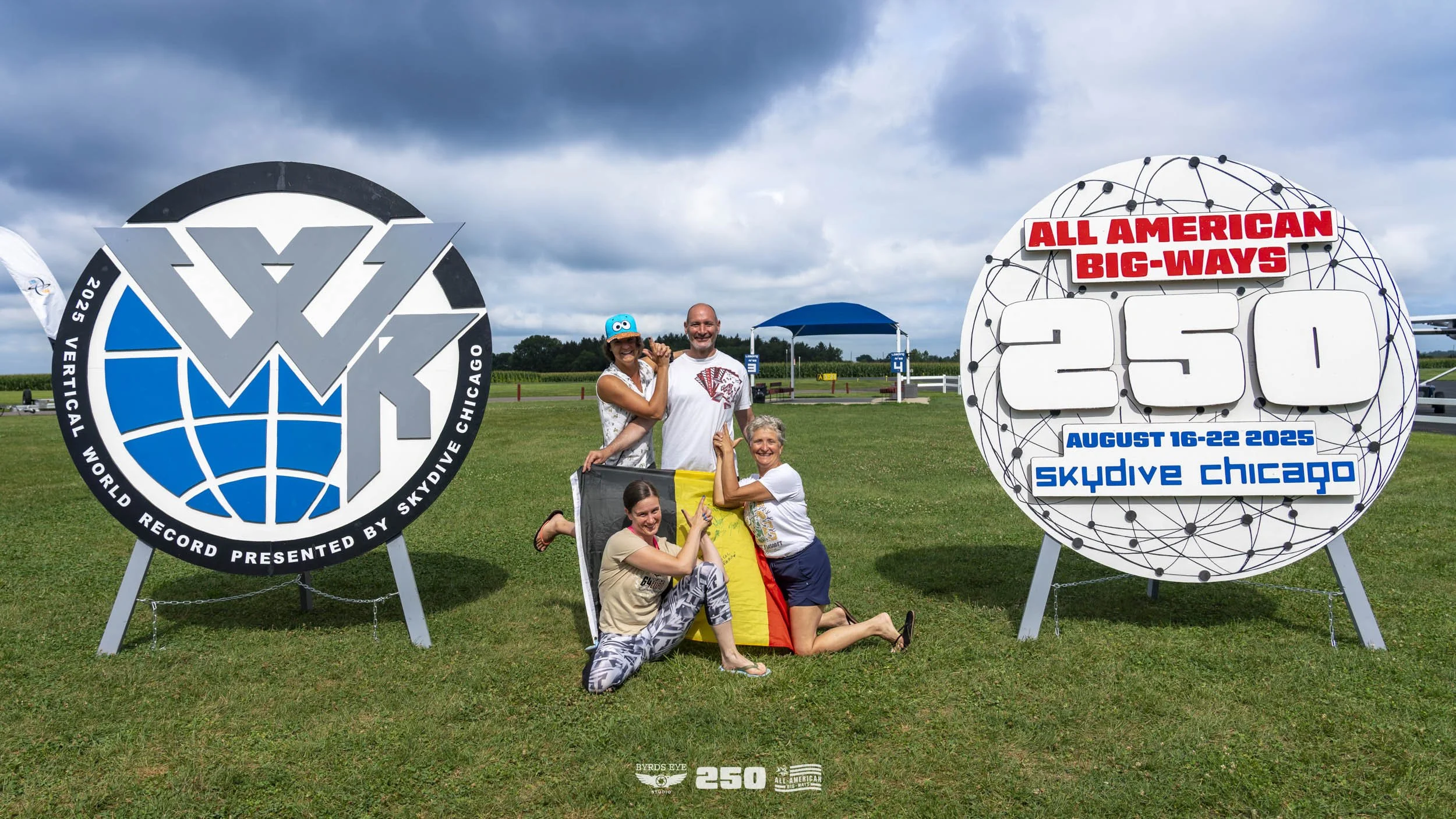 Group of five people posing on grass next to sign celebrating a Guinness World Record for the largest indoor skydiving flight, with one person holding a German flag. Signs display event details: All American Big-Ways, August 16-22, 2025, Skydive Chic