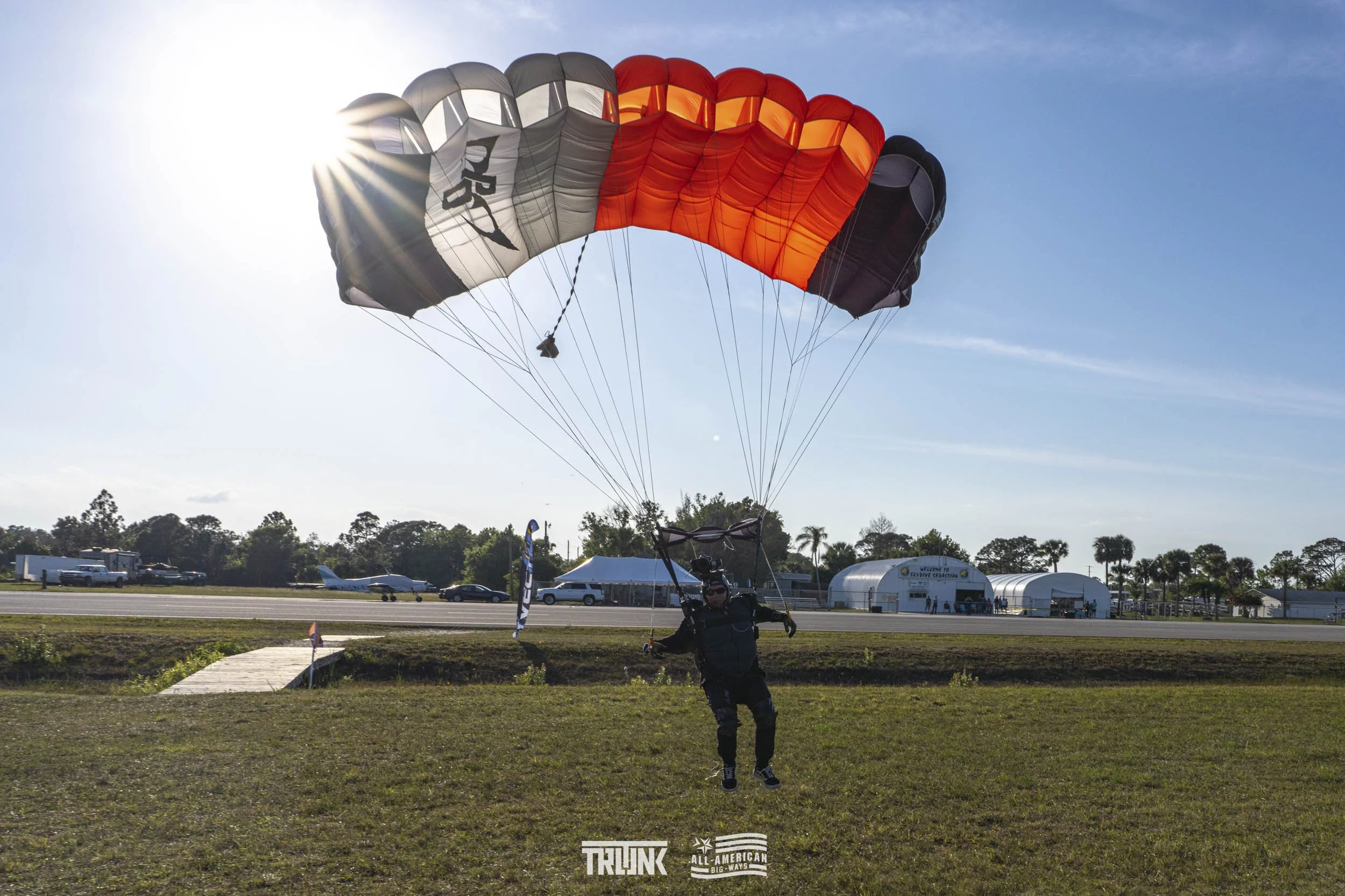 Man landing with a parachute on a grassy field near an airstrip with planes and hangars in the background.