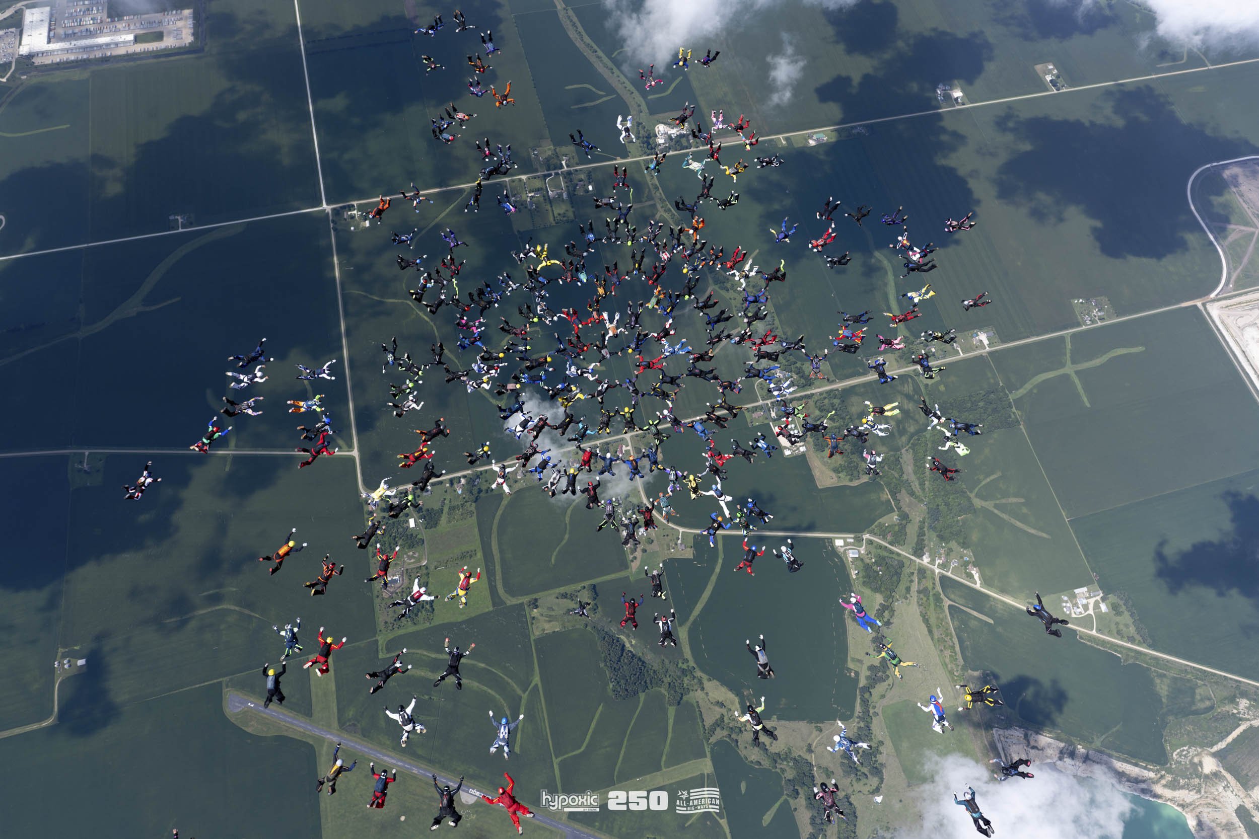 A large group of skydivers in colorful jumpsuits, mid-air during a skydiving formation over a landscape with fields, roads, and clouds.