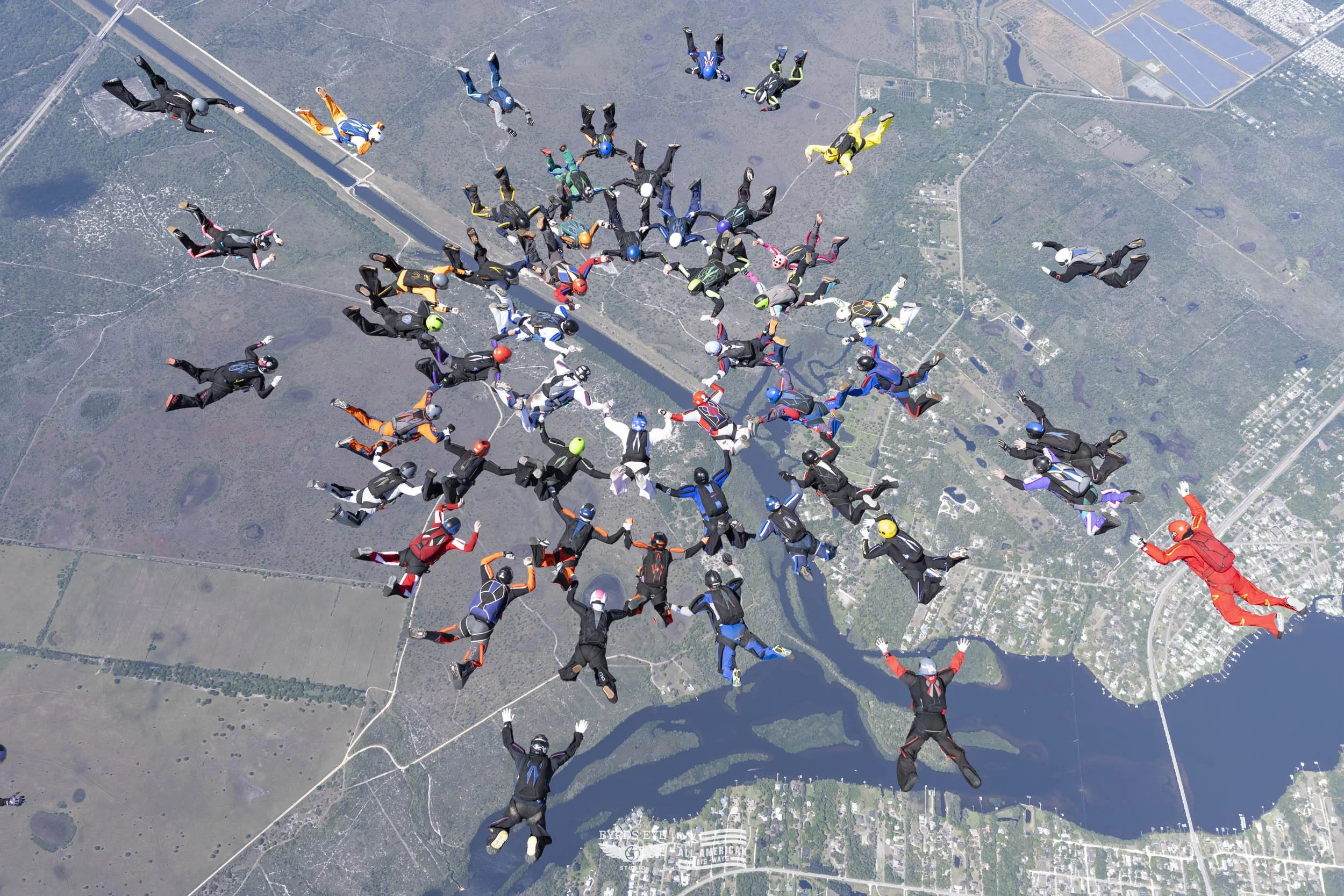 Group of skydivers in free fall above a landscape with water bodies, roads, and greenery.