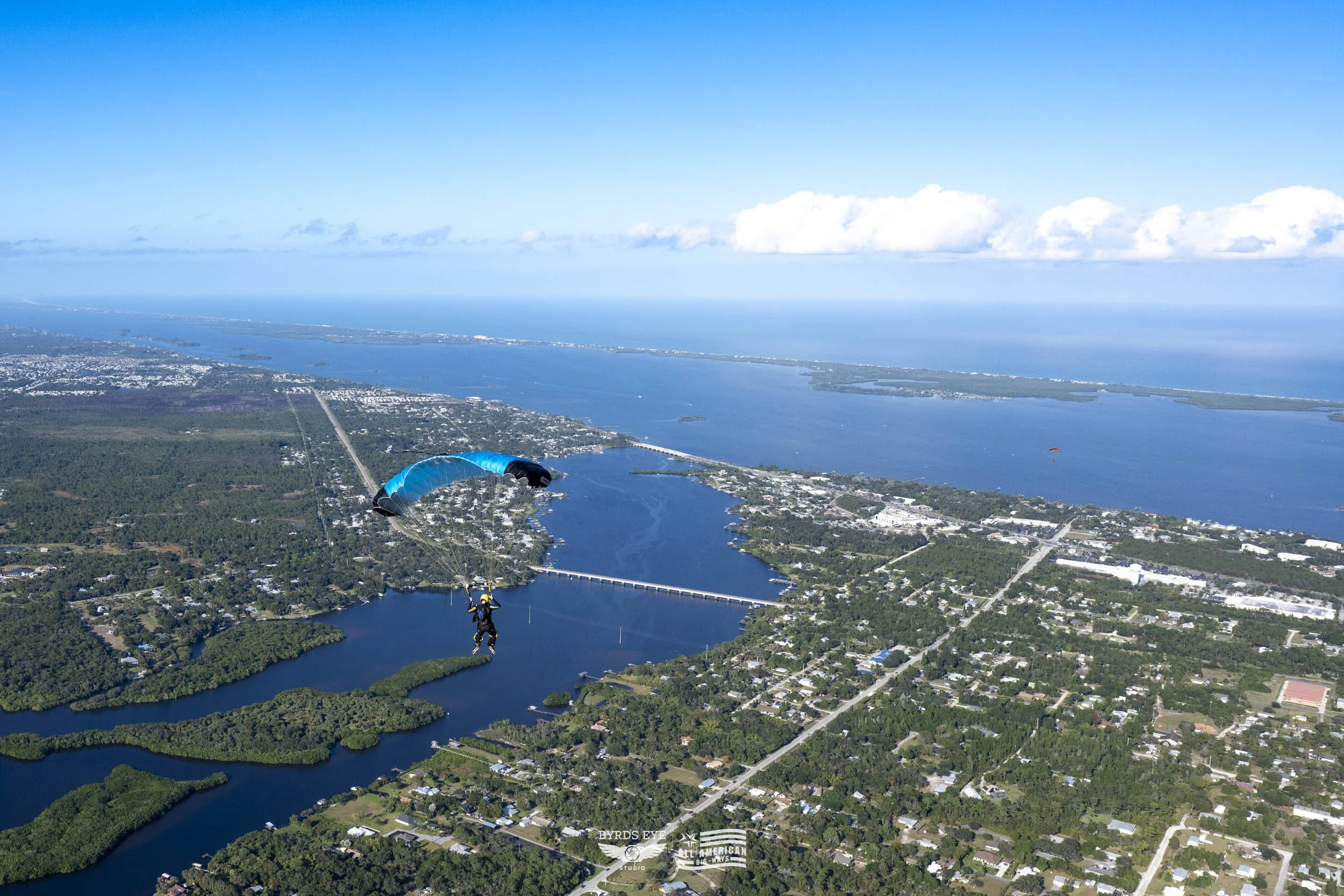 Person tandem skydiving over a city and water bodies, with a blue sky and clouds in the background.
