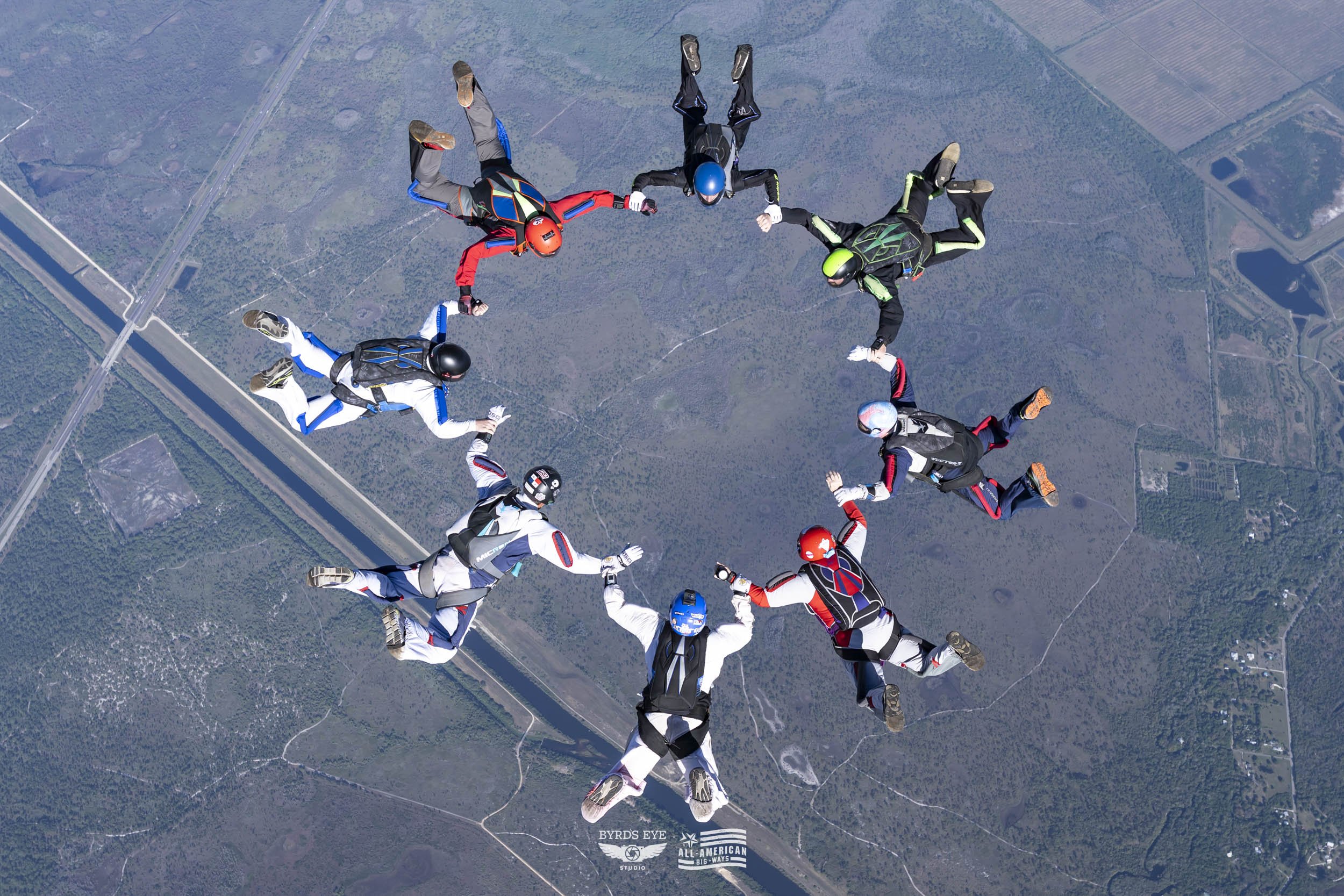 A group of eleven skydivers holding hands in a circle while free-falling over a landscape with fields, roads, and a river.
