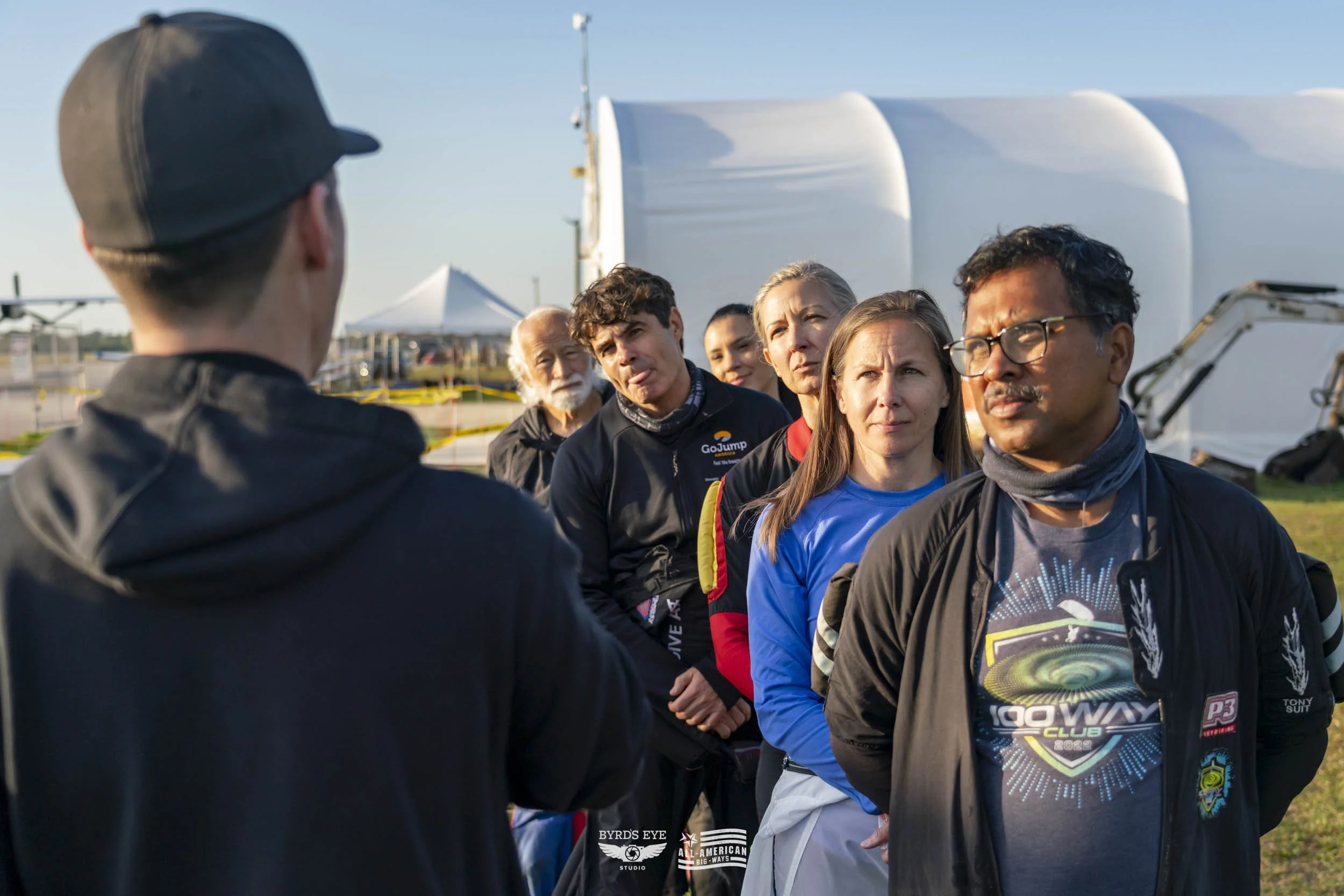 A group of people standing outdoors, listening to a person in a hat speaking.