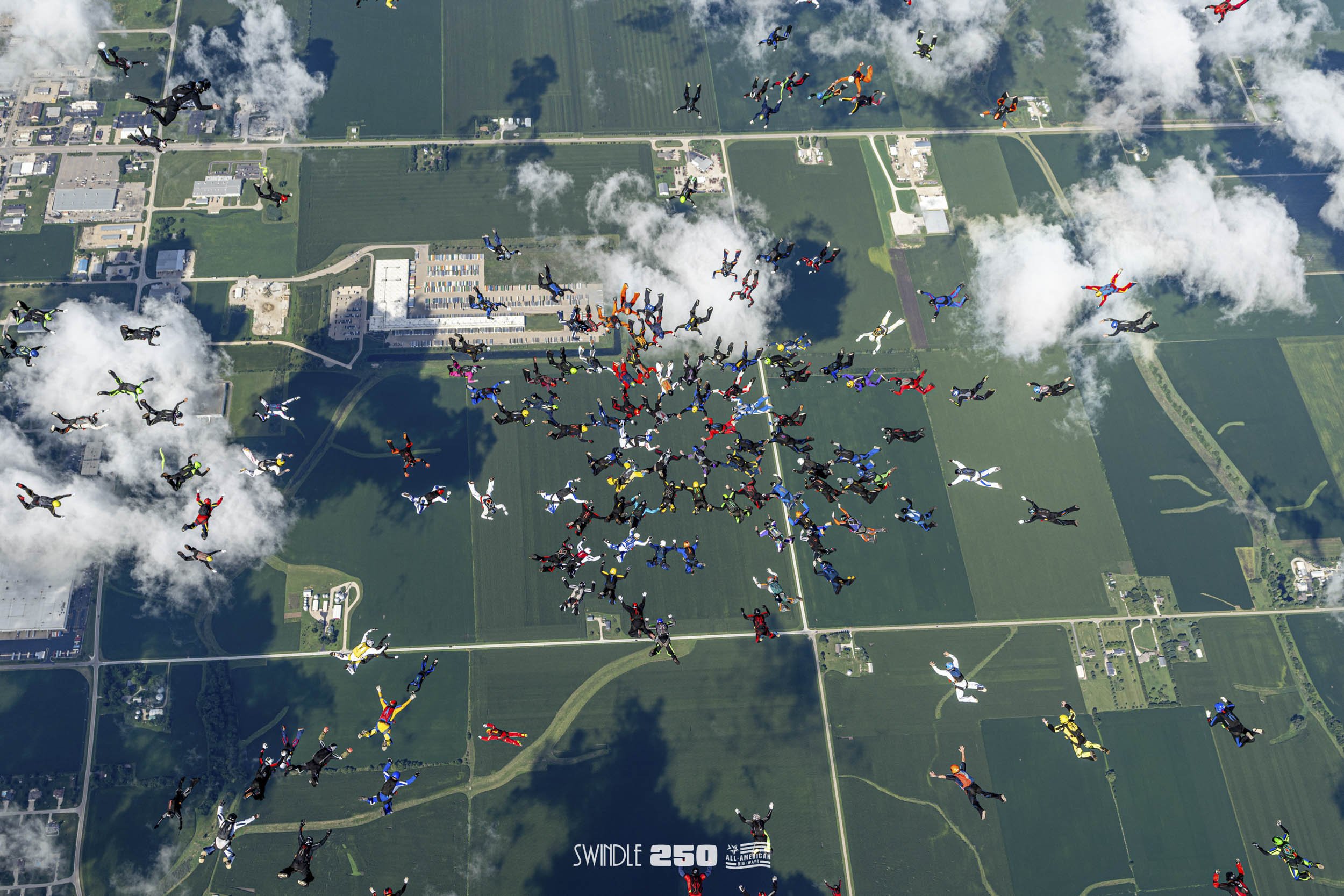 An aerial view of hundreds of skydivers in colorful jumpsuits and parachutes in free fall over a green landscape with fields and roads.