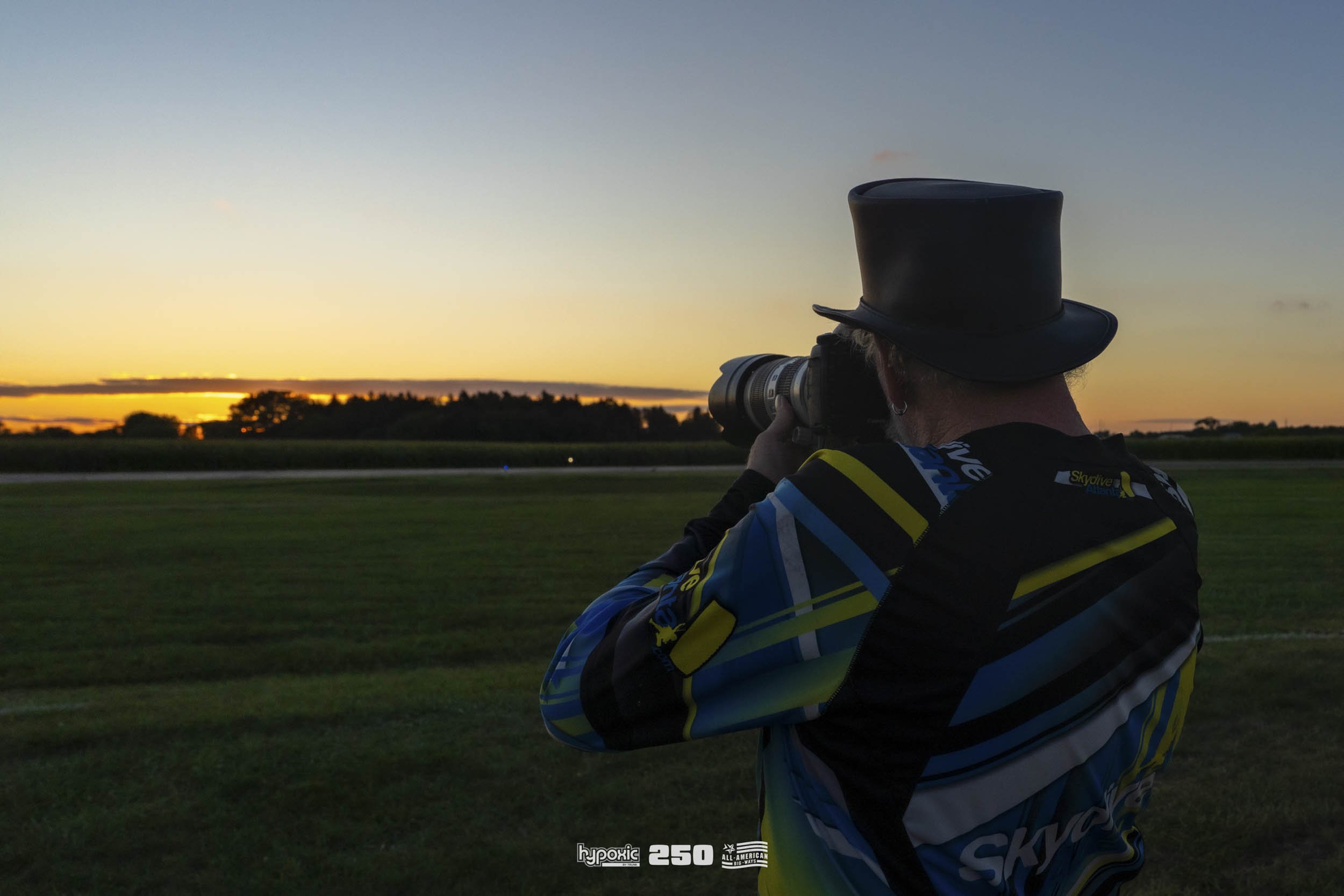 Person wearing a top hat and colorful sports jersey is aiming a camera at a sunset over a grassy field, with trees in the background.