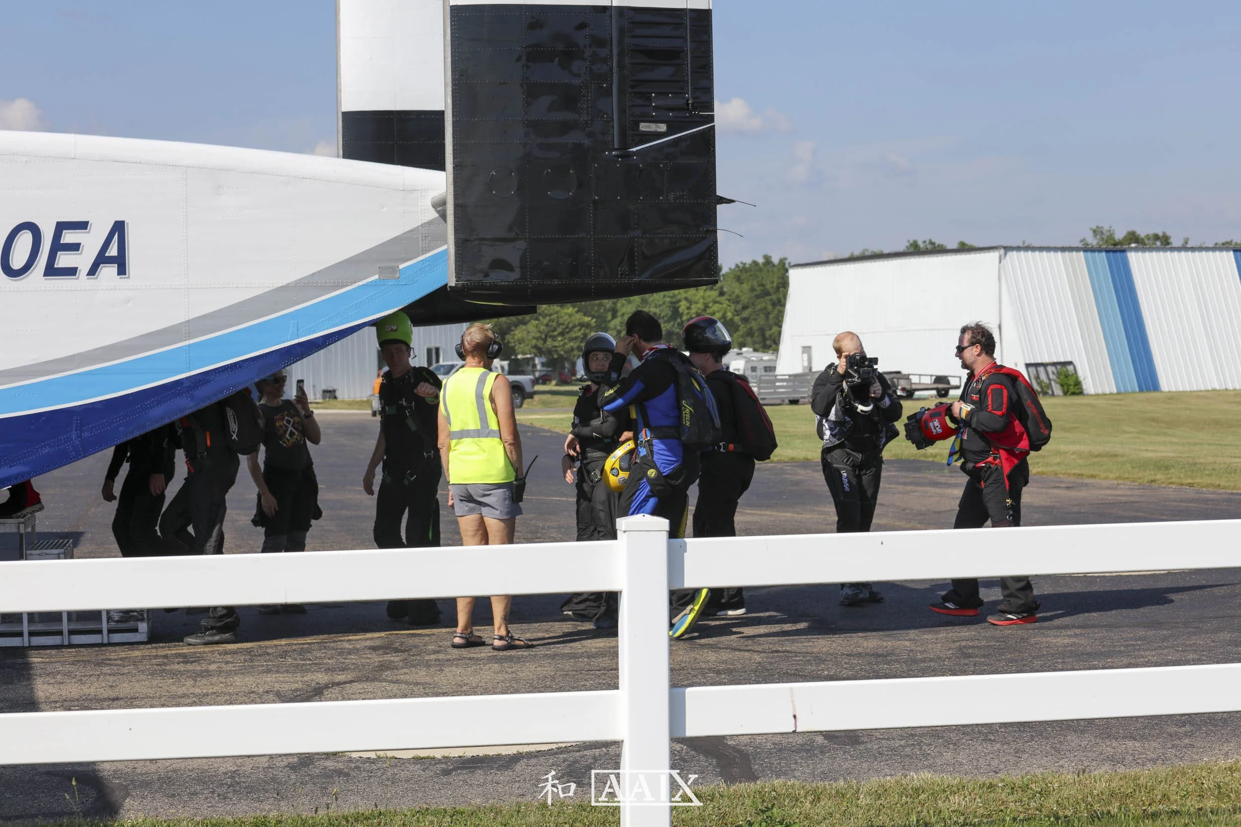 A group of skydivers in jumpsuits and helmets standing next to a large aircraft on the tarmac, preparing for a jump with some members talking and others taking photos.