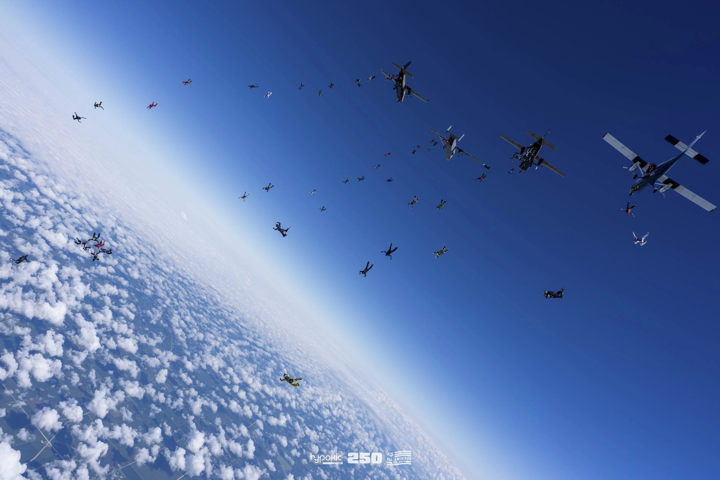 Skydivers in freefall formation with planes flying overhead, seen from above the clouds.