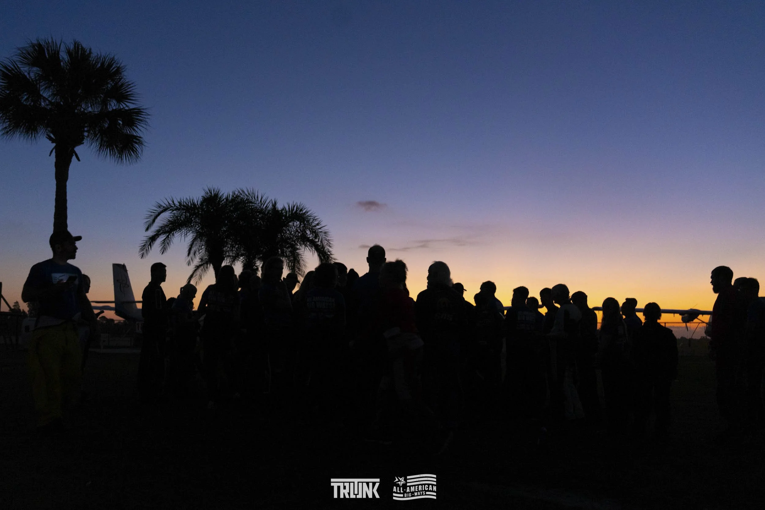Silhouetted group of people and palm trees against a colorful sunset sky.