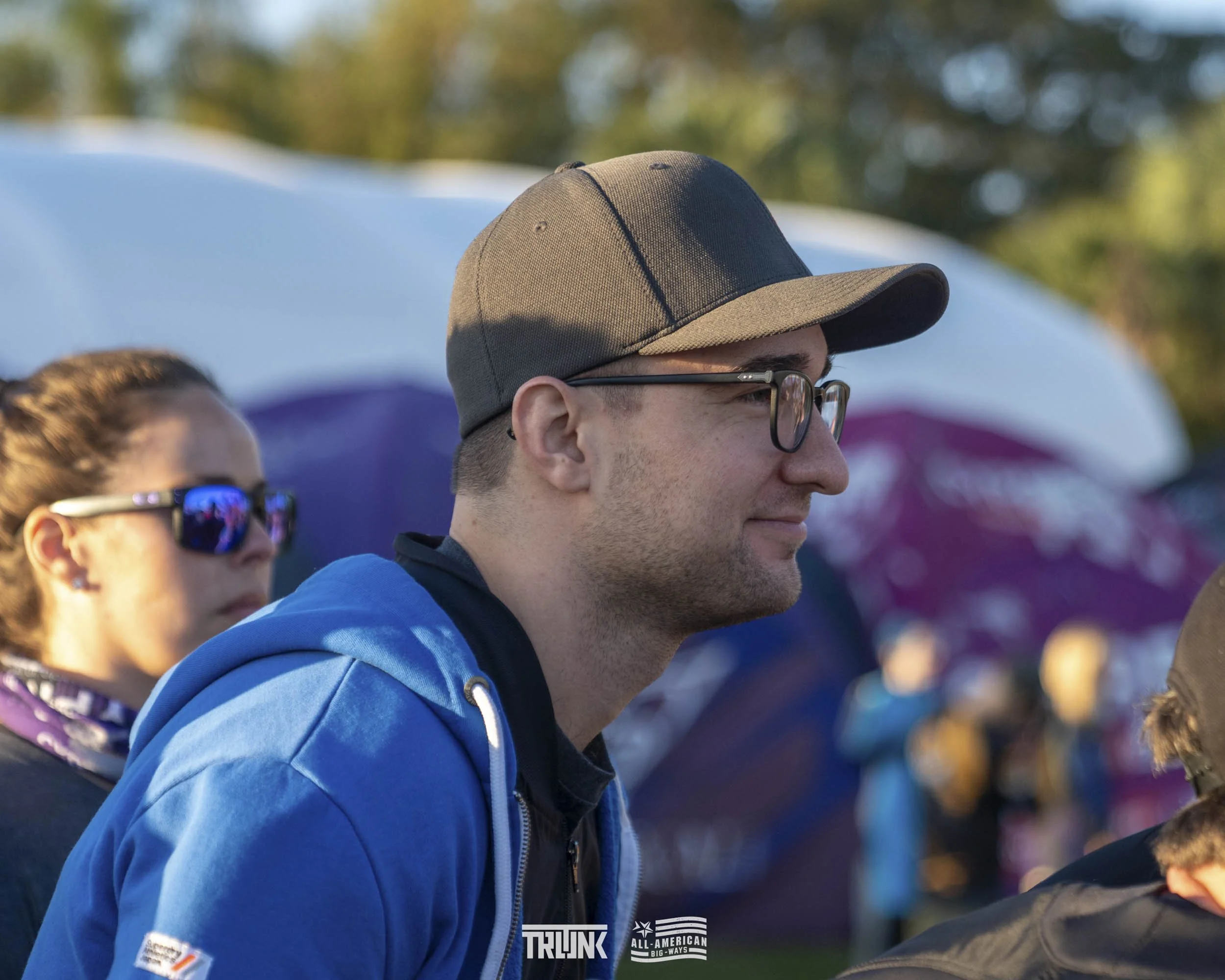 A man in a gray baseball cap, glasses, and a blue hoodie, standing outdoors at dusk with trees and tents in the background.