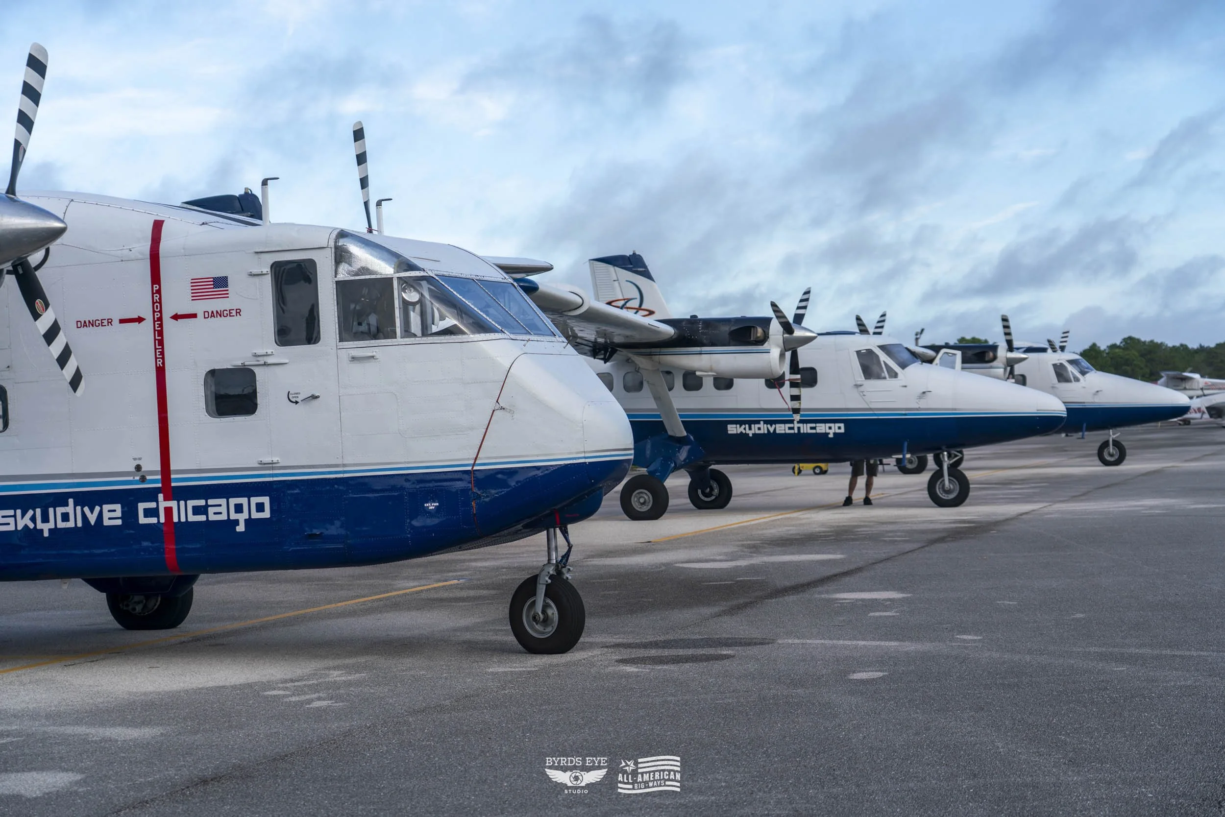 Several small aircraft parked on an airstrip, with people walking nearby. The aircraft are white and blue, one labeled 'skydive chicago'.