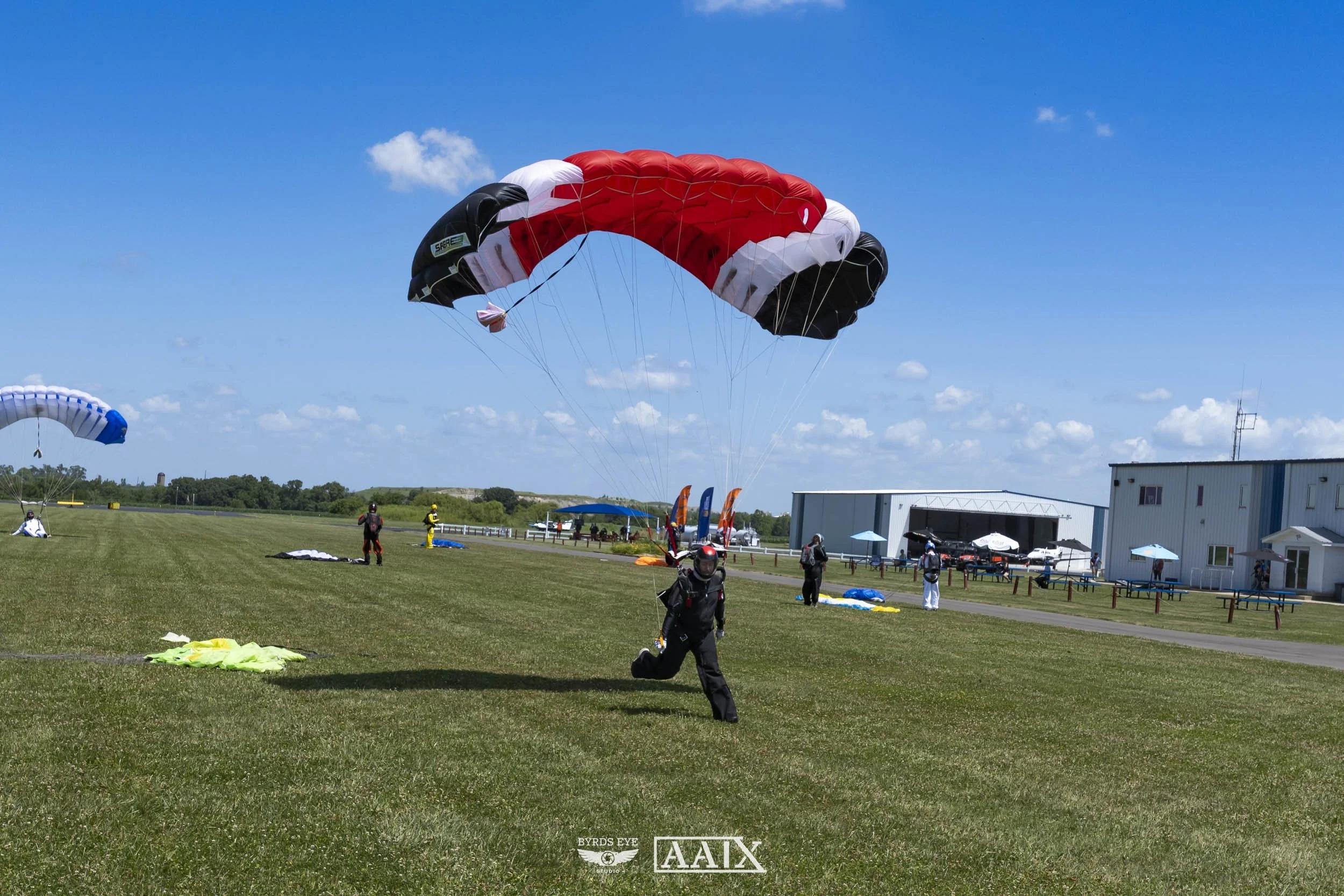 A person running on a grassy field after landing from a tandem skydiving jump with a red, white, and black parachute. Other parachutes and skydivers are visible on the ground and in the air, with a hangar and blue sky in the background.