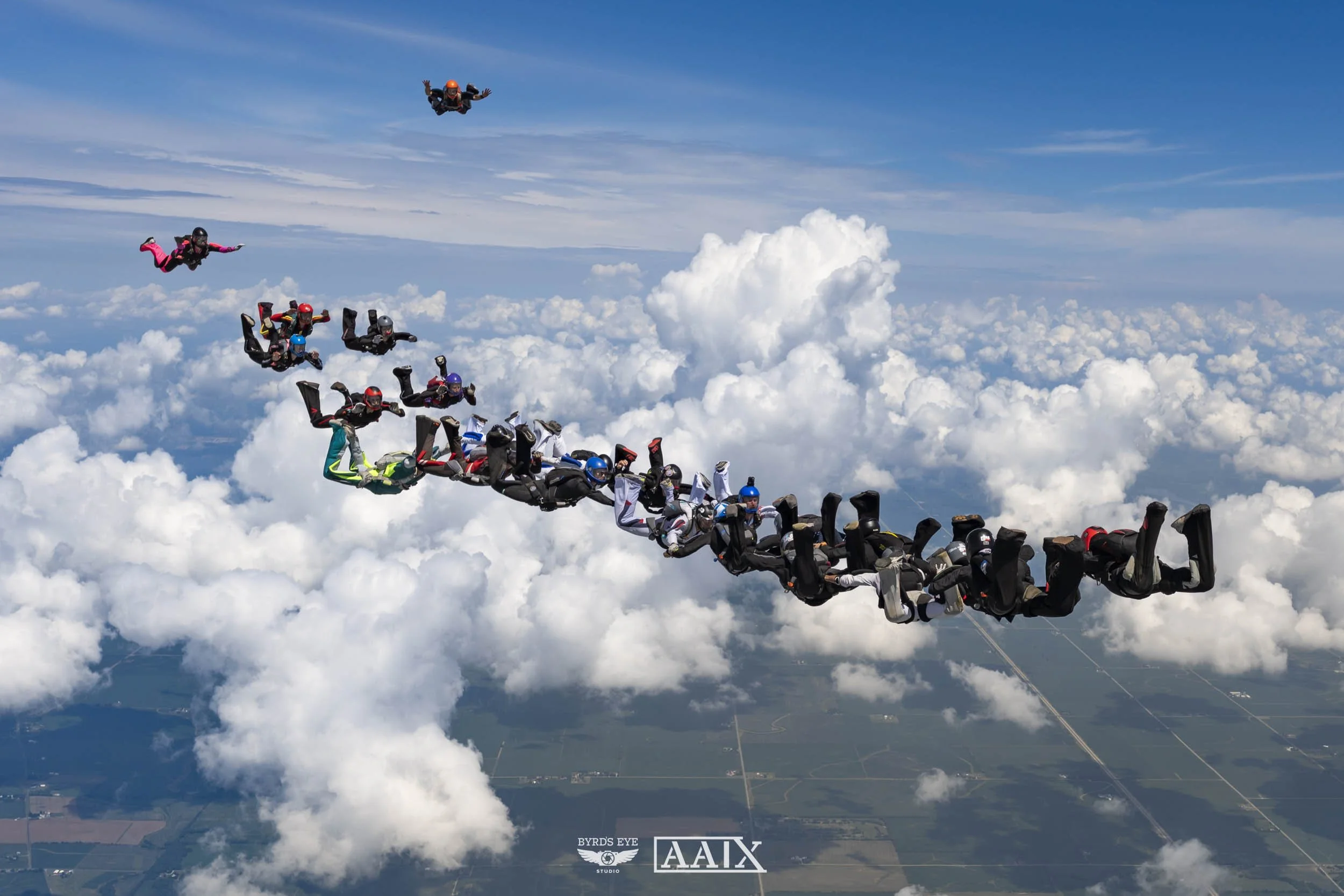A group of skydivers in free fall above the clouds, with blue sky and clouds in the background.