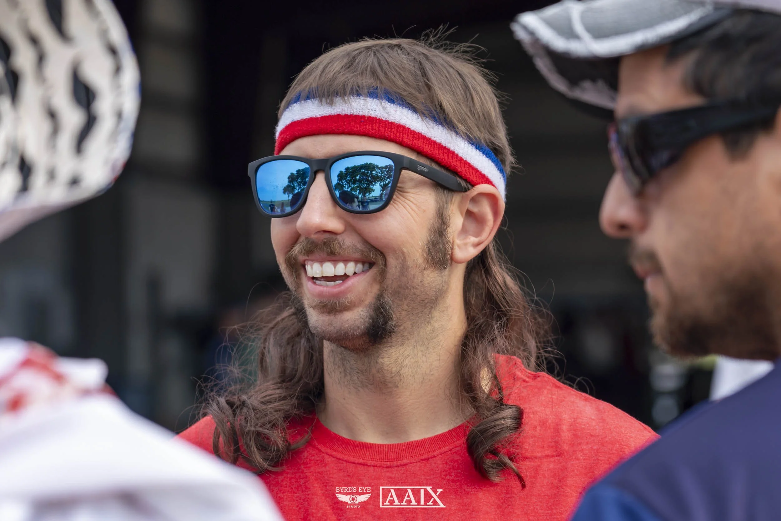 A man with long hair and a beard smiling, wearing sunglasses, a red shirt, and a red, white, and blue headband, engaged in conversation with two other men.