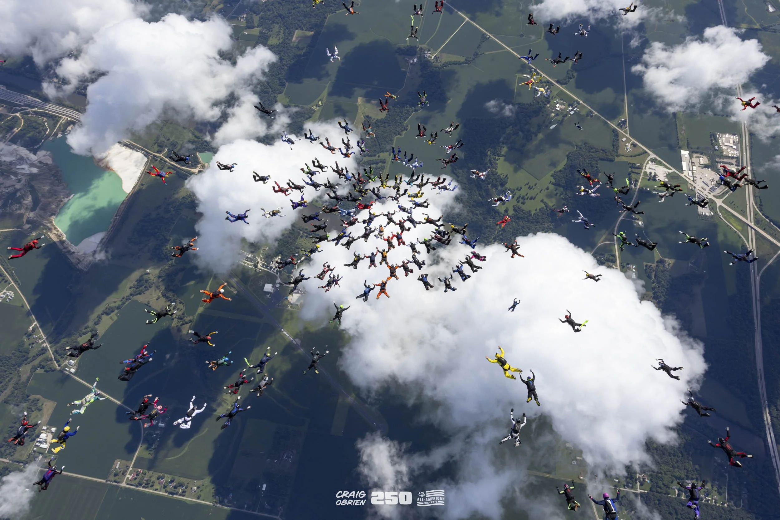Skydivers in colorful jumpsuits free-falling from above, forming circular patterns among the clouds over a landscape with lakes, forests, and roads.
