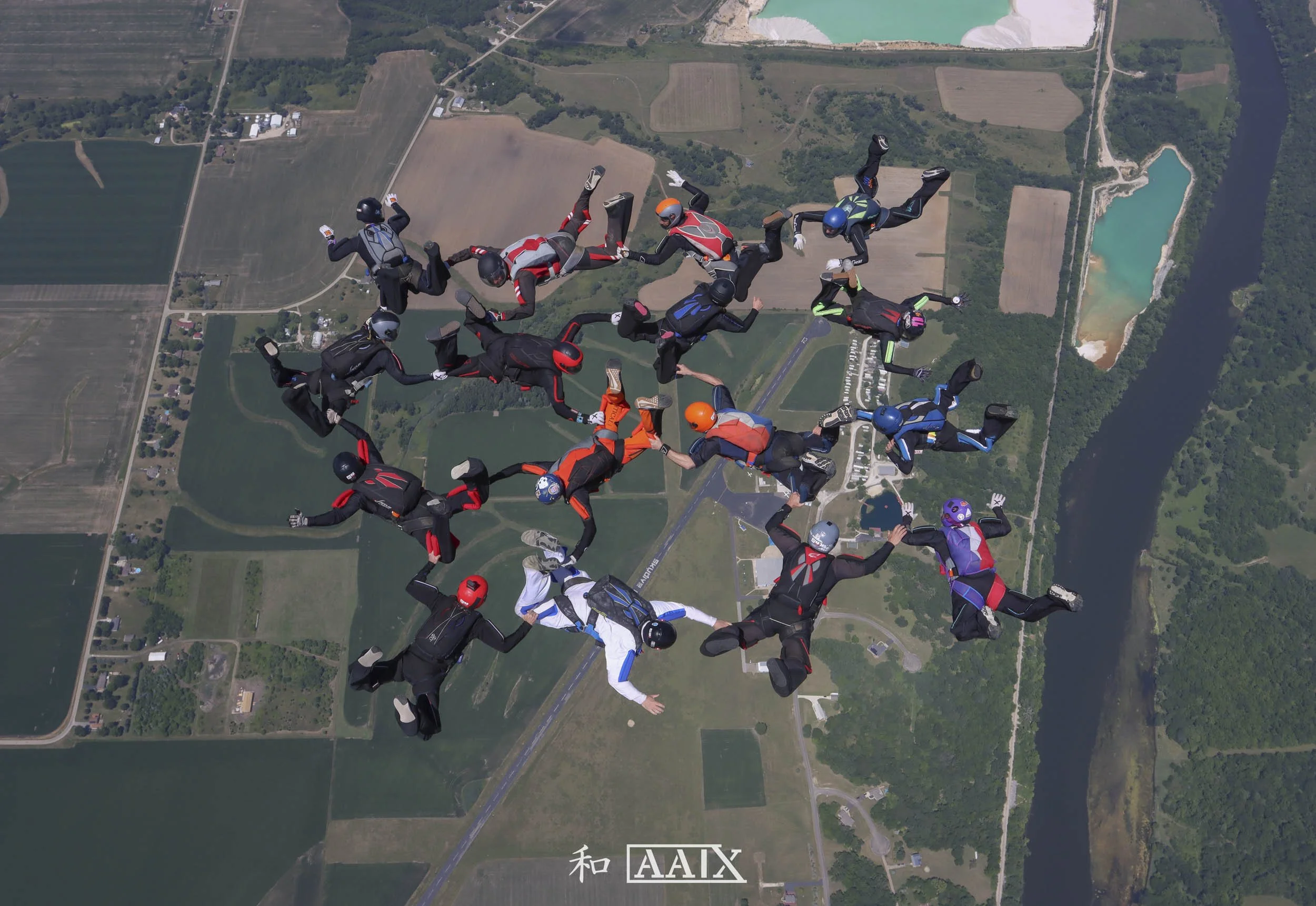 Group of skydivers holding hands while free-falling over a rural area with fields, a river, and a lake.