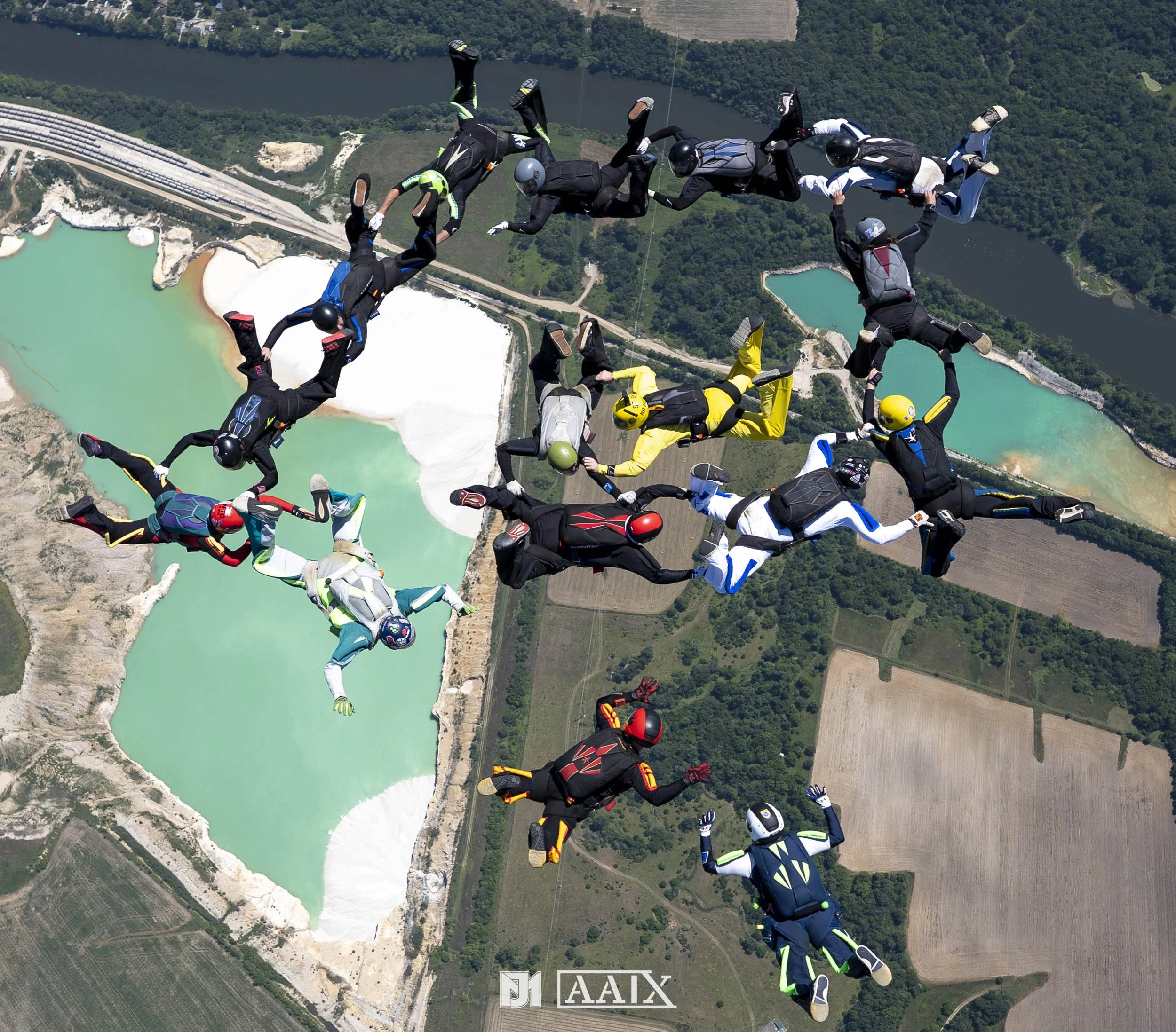 A group of skydivers free-falling over a landscape with lakes, forests, and farmland, forming a circle with their bodies and holding hands.